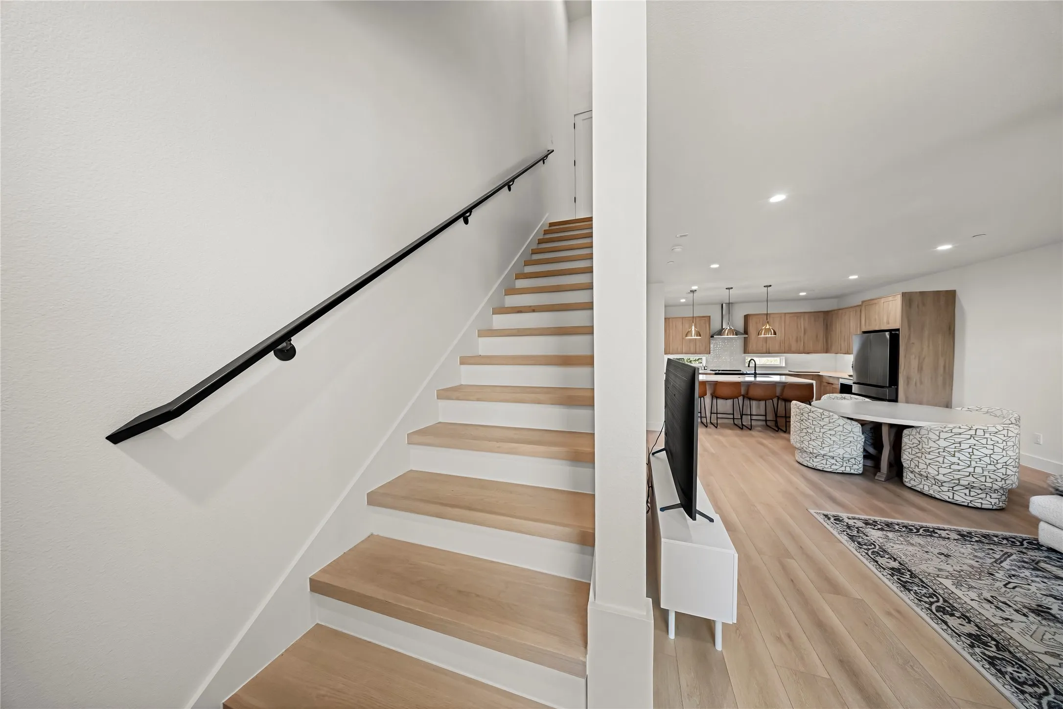 Light wood-finish flooring extending into the kitchen, open-concept living area, modern kitchen cabinetry, black appliances, and a straight staircase with a black handrail