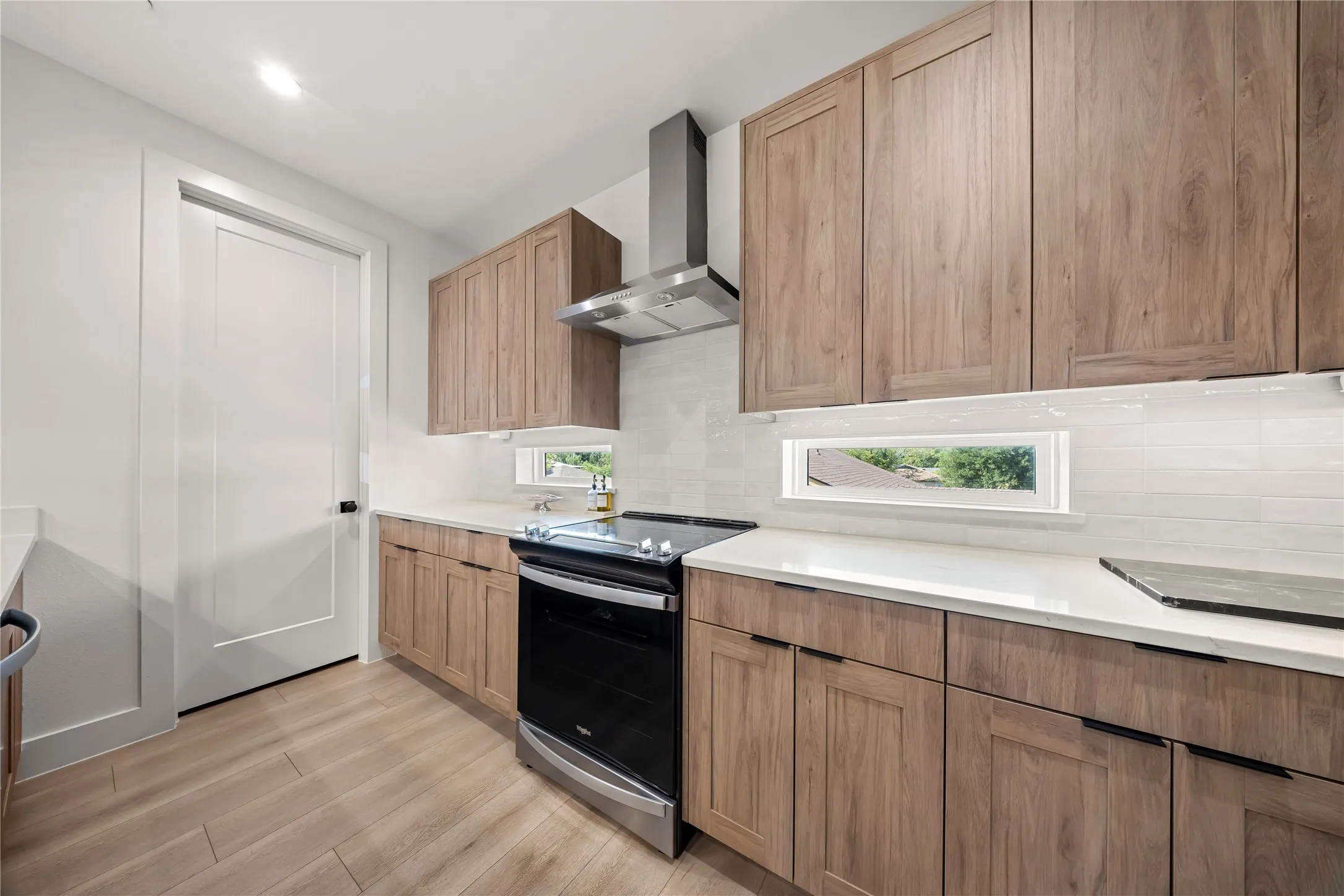 Kitchen featuring wood-finish cabinetry, light-toned countertops, a stainless steel range hood, white subway tile backsplash, and wood-finish flooring