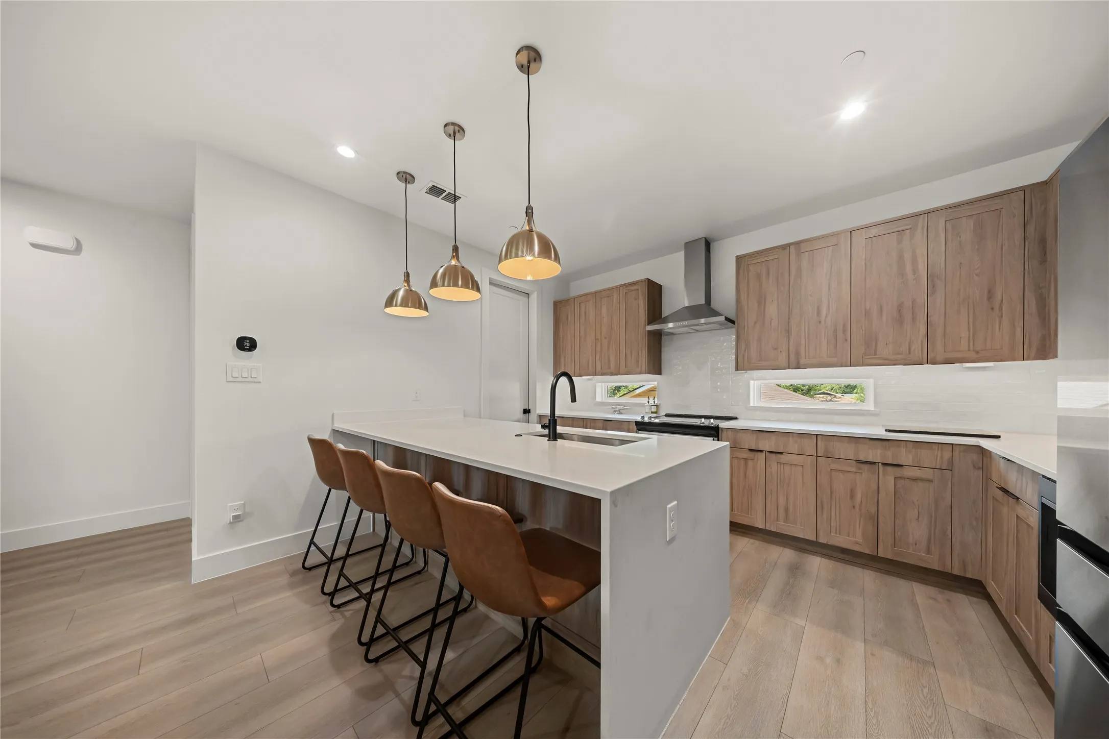 Kitchen featuring wood-finish cabinetry, stainless steel range hood, light wood-finish flooring, white countertops, and three brass-finish pendant lights