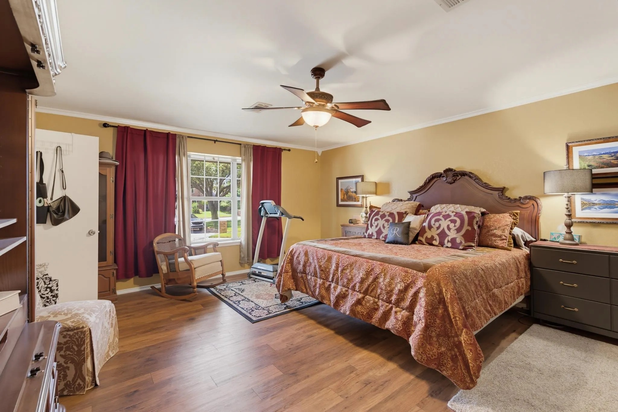 Spacious primary bedroom suite featuring hardwood flooring, a ceiling fan with integrated lighting, arched headboard bed frame, and large windows with burgundy drapery