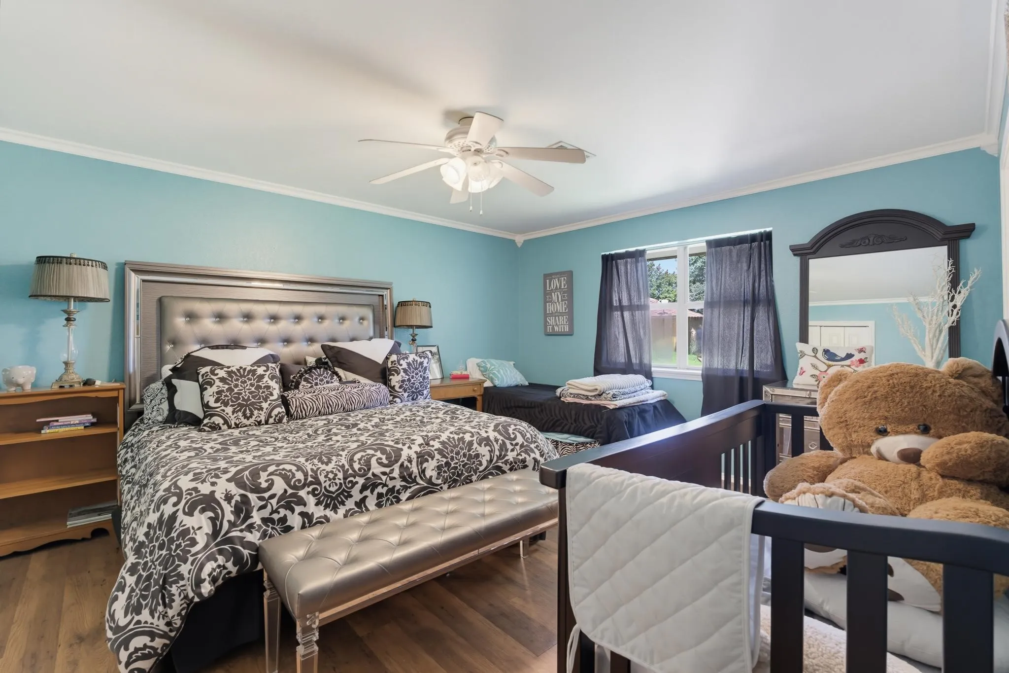 Bright bedroom retreat showcasing turquoise accent walls, a tufted upholstered headboard with nailhead detailing, ceiling fan with integrated lighting, wood-finish flooring, and a crib positioned within the room
