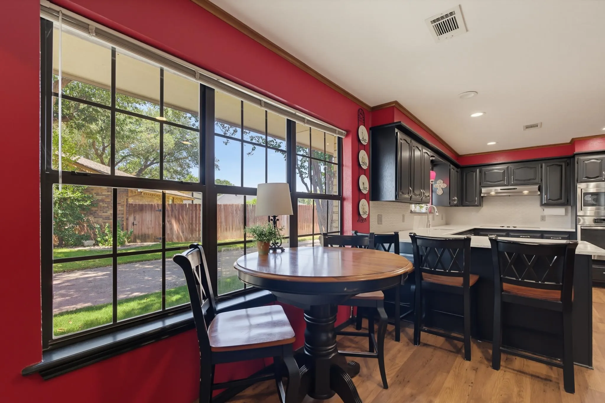 Bright dining area with bold crimson accent walls, large multi-pane windows framing outdoor scenery, wood-finish flooring, and black cabinetry with integrated appliances visible in the adjoining kitchen space