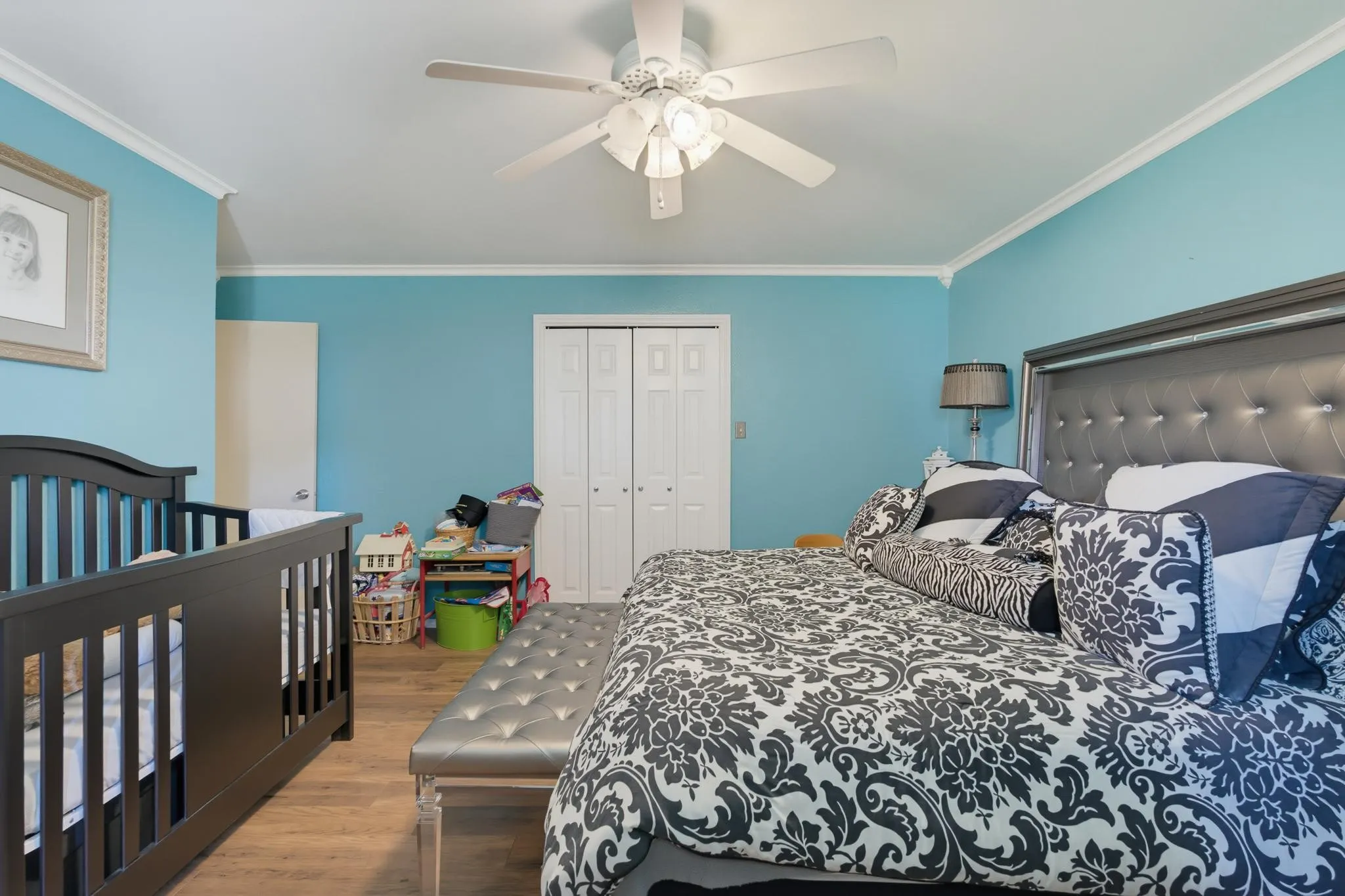 Spacious master bedroom suite featuring soft turquoise walls, ceiling fan with integrated lighting, wood-finish flooring, and generous square footage accommodating a full-size bed and separate crib