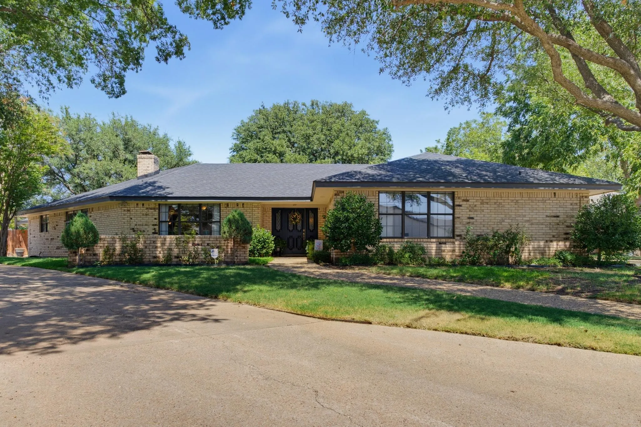 Brick exterior with a dark-shingled roof, mature trees, and a concrete driveway