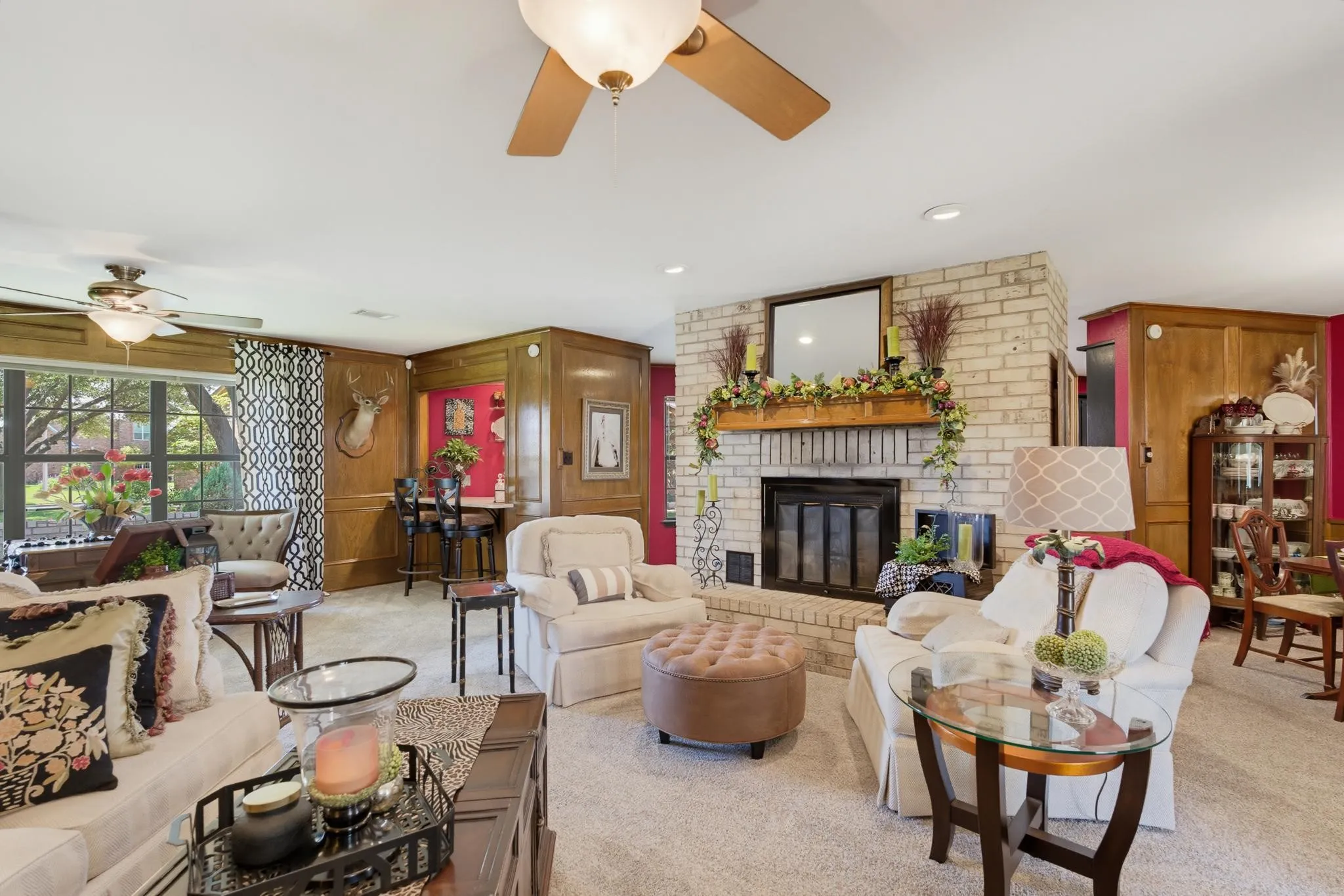 Expansive living area featuring a brick fireplace with a wood mantle, broad window with exterior landscaping, built-in wood paneling, and two ceiling fans