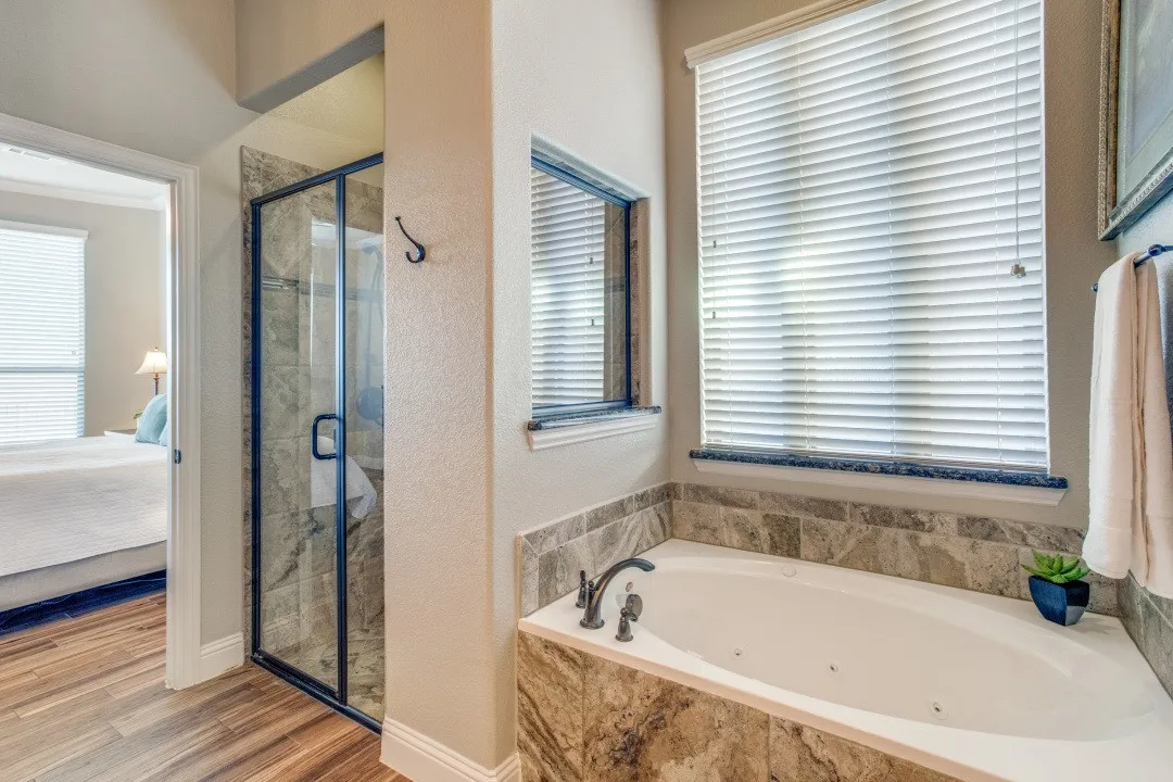 Spa-inspired primary bathroom featuring a soaking tub with surround, walk-in shower with frameless glass enclosure and tile surround, dual vanity with stone countertop, horizontal blinds at large windows, and wood-finish flooring throughout