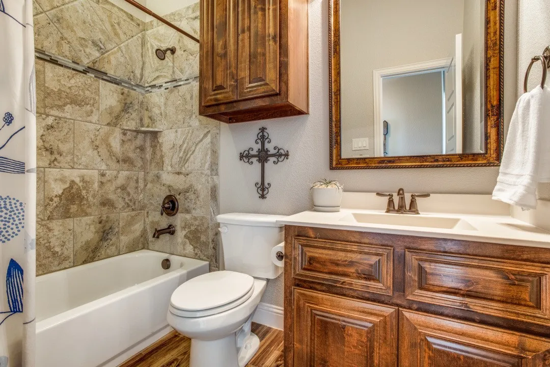 Wood-finish flooring, a wood-finish vanity with an integrated sink, a framed mirror, and a tiled shower with a built-in shelf