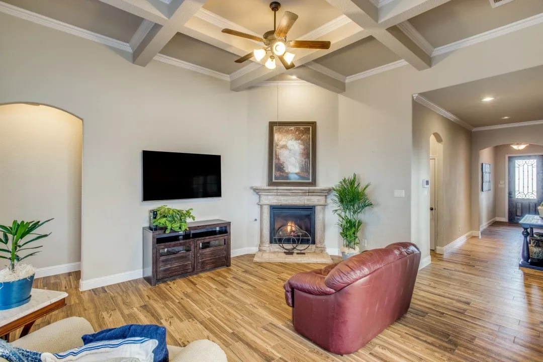 Vaulted ceiling with decorative beams, wood-finish flooring, corner fireplace with stone surround, ceiling fan, and arched entryway