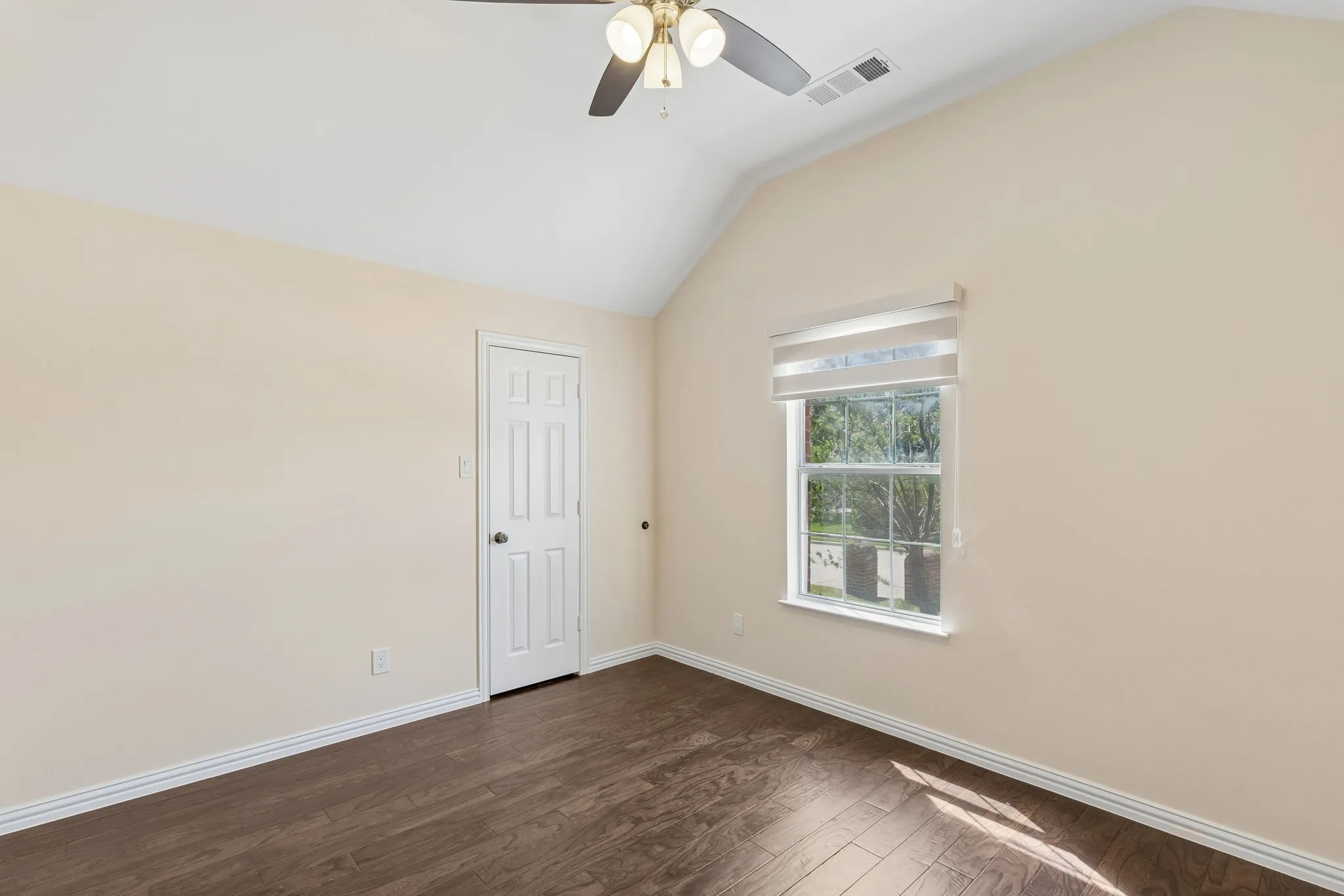 Vaulted ceiling, wood-finish flooring, ceiling fan with integrated lighting, window with roller shade, and paneled interior door