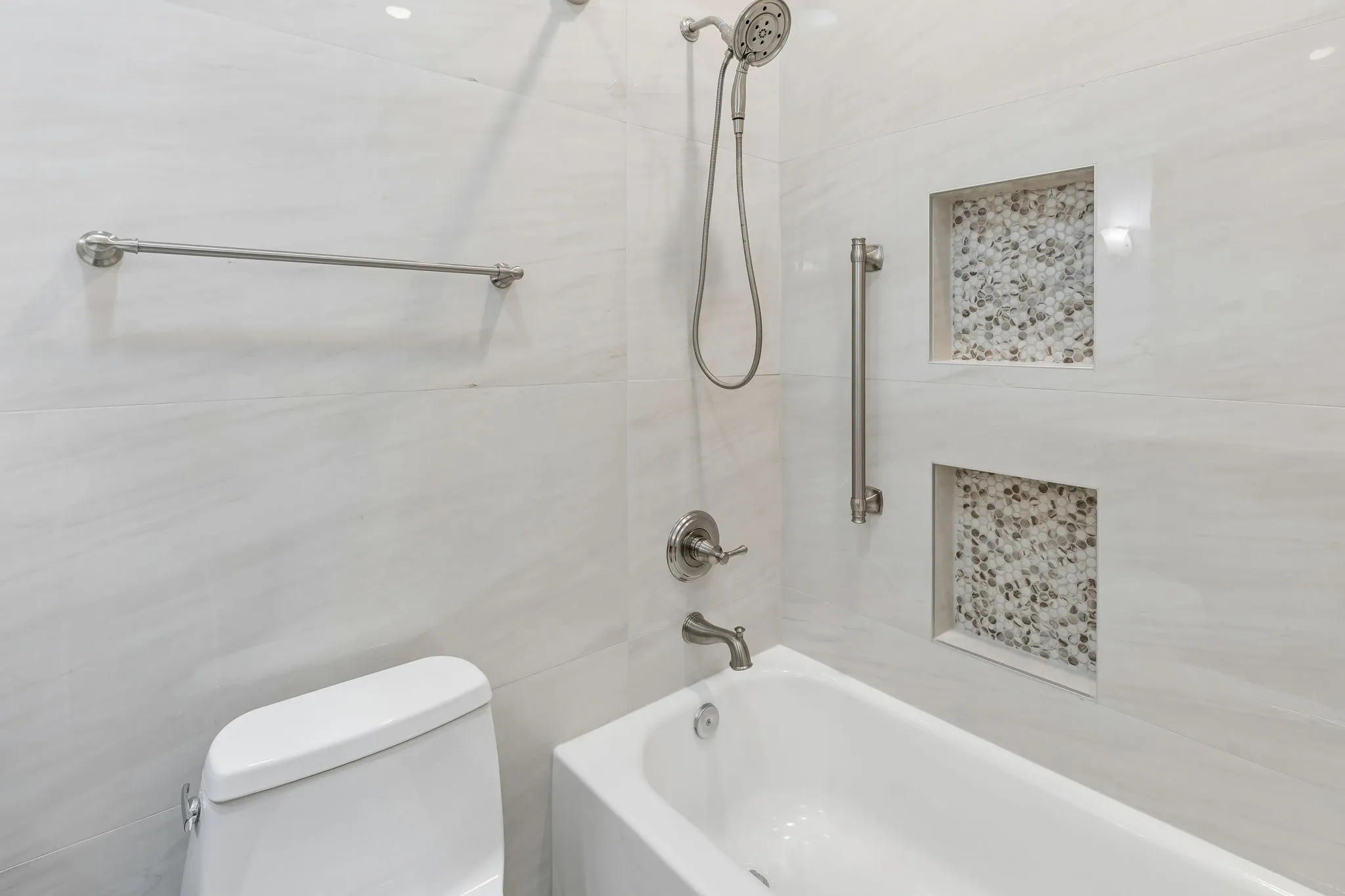 Modern bathroom featuring crisp white subway tile walls, a soaking tub with integrated deck-mounted faucet and handheld showerhead, polished chrome fixtures, recessed shelving with decorative mosaic tile inserts, and coordinating towel bar hardware