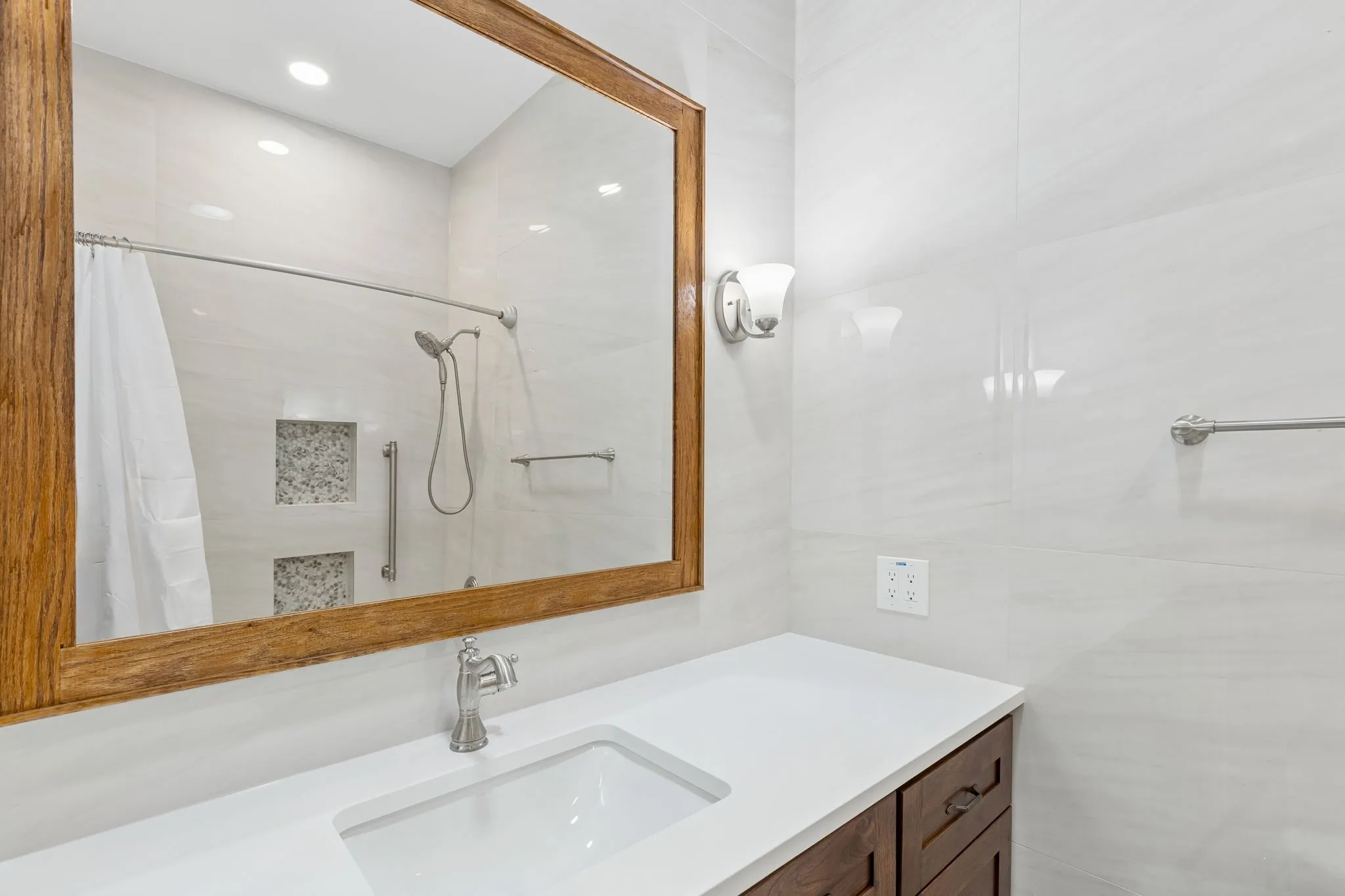 Quartz countertop and undermount sink, wood-finish cabinetry, a large wood-framed mirror, white wall tiling, and sconce lighting. Tile niches in shower and heated floors!