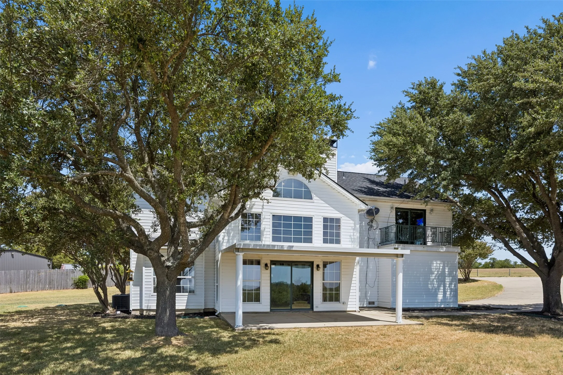 White exterior siding with a covered patio, upper-level balcony, arched window, and mature trees