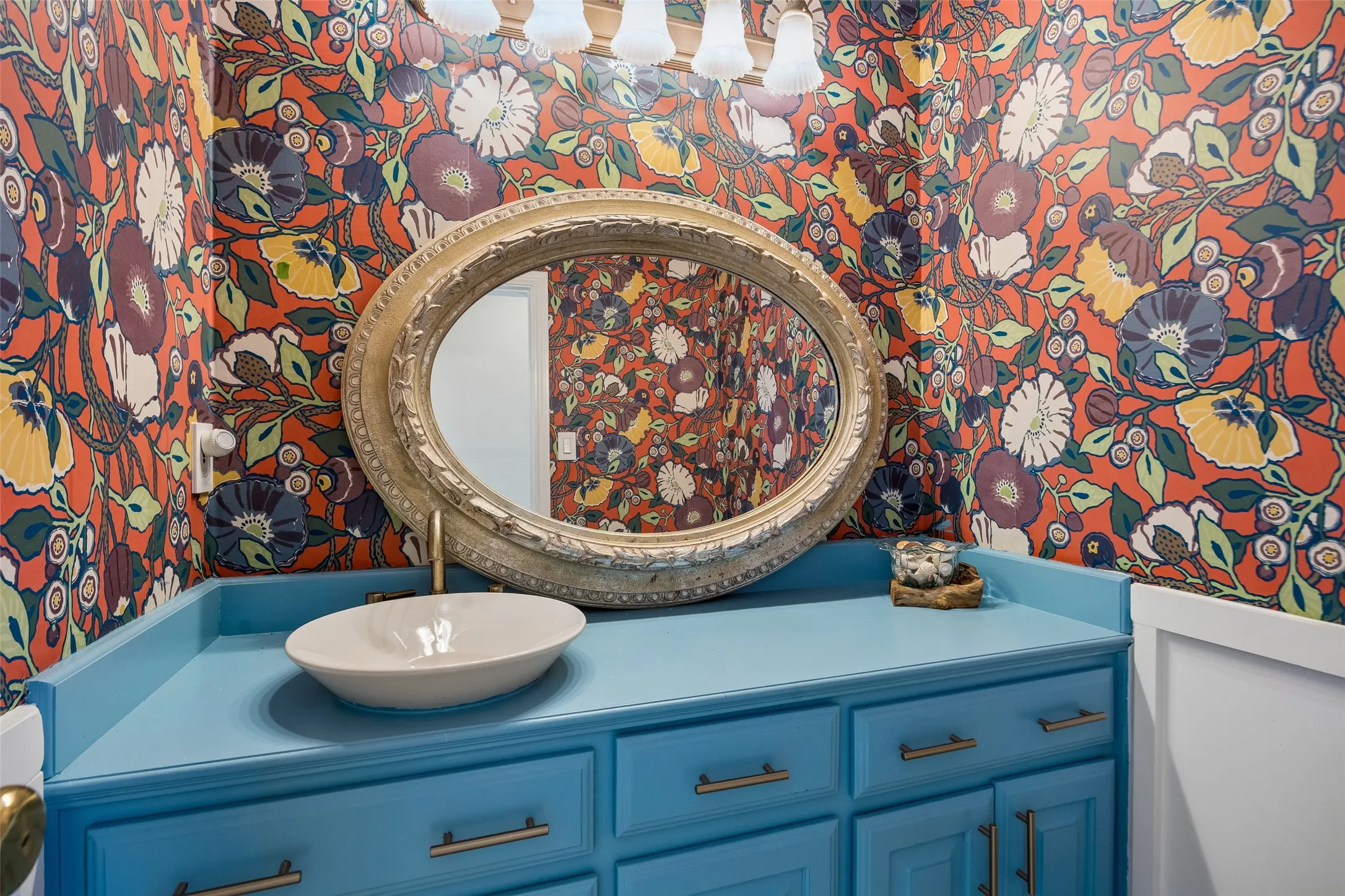 First floor powder bath Colorful floral wallpaper, oval vanity mirror, vessel sink, blue cabinetry, and wall-mounted light fixture