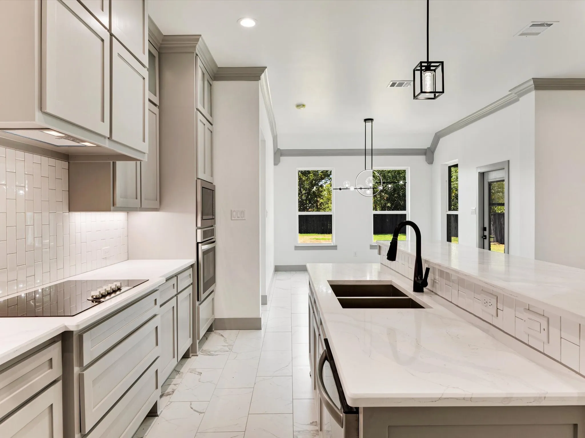Modern kitchen featuring light gray cabinetry, white countertops, stainless steel double wall ovens, a smooth cooktop, and a kitchen island with a double basin sink and matte black faucet