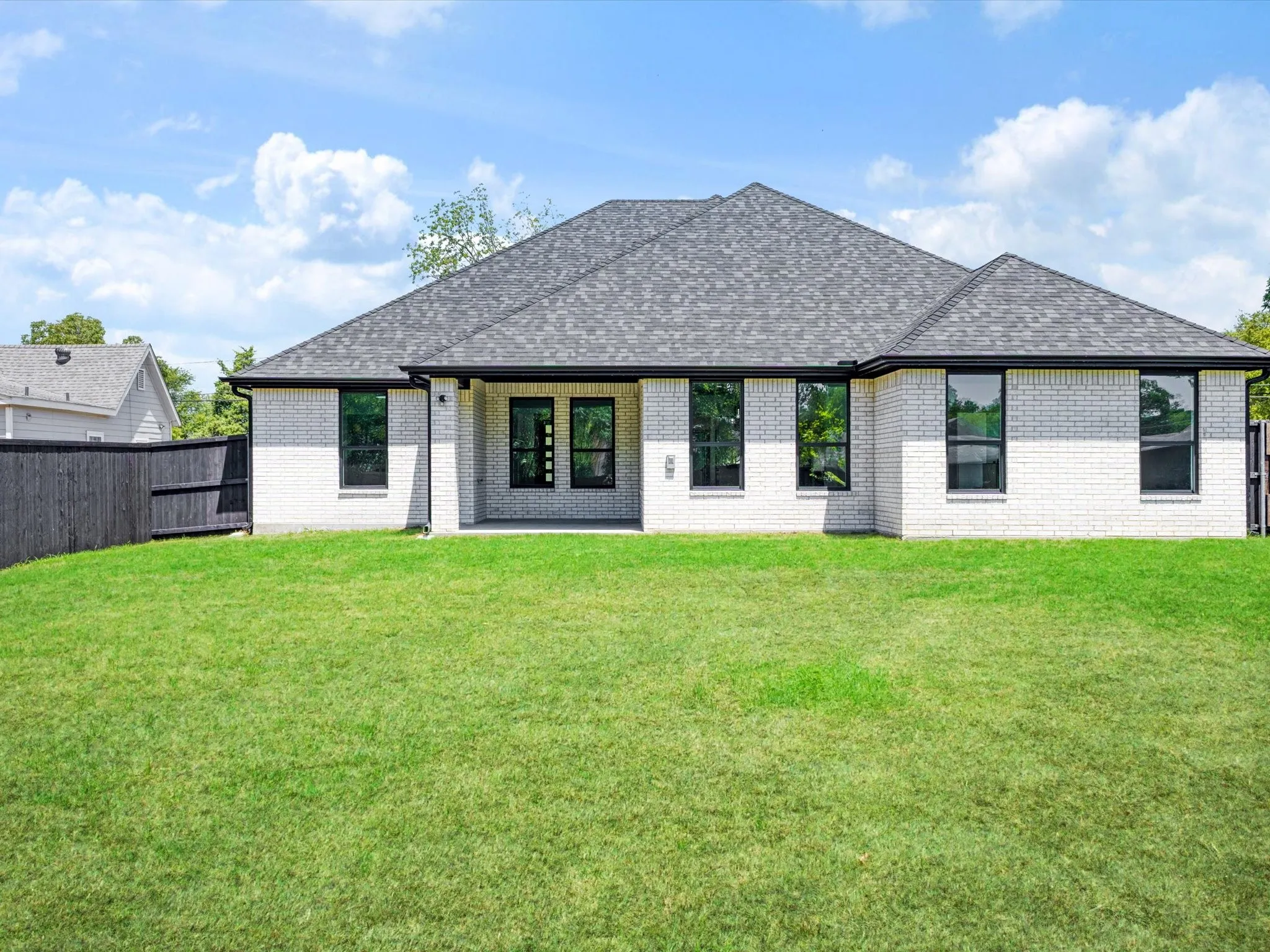 White brick exterior, dark grey shingle roof, black privacy fence, rear patio, and expansive green lawn