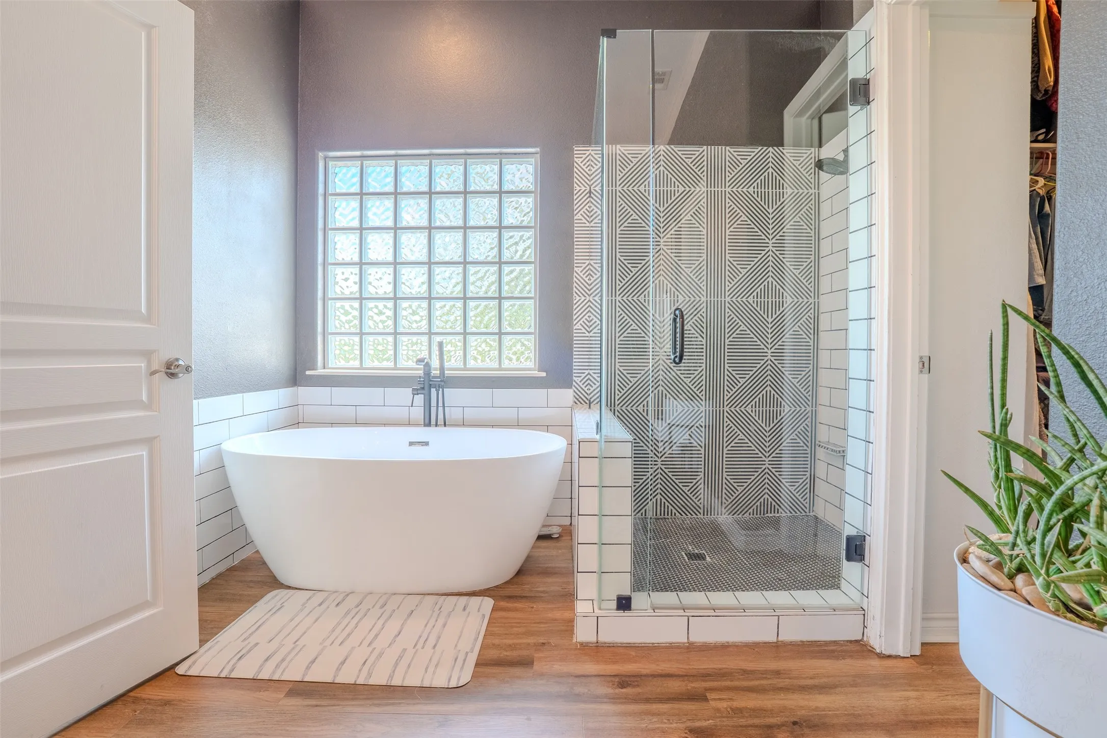 Freestanding soaking tub with chrome fixtures, glass block window, frameless glass shower enclosure, decorative patterned shower wall tile, and wood-finish flooring