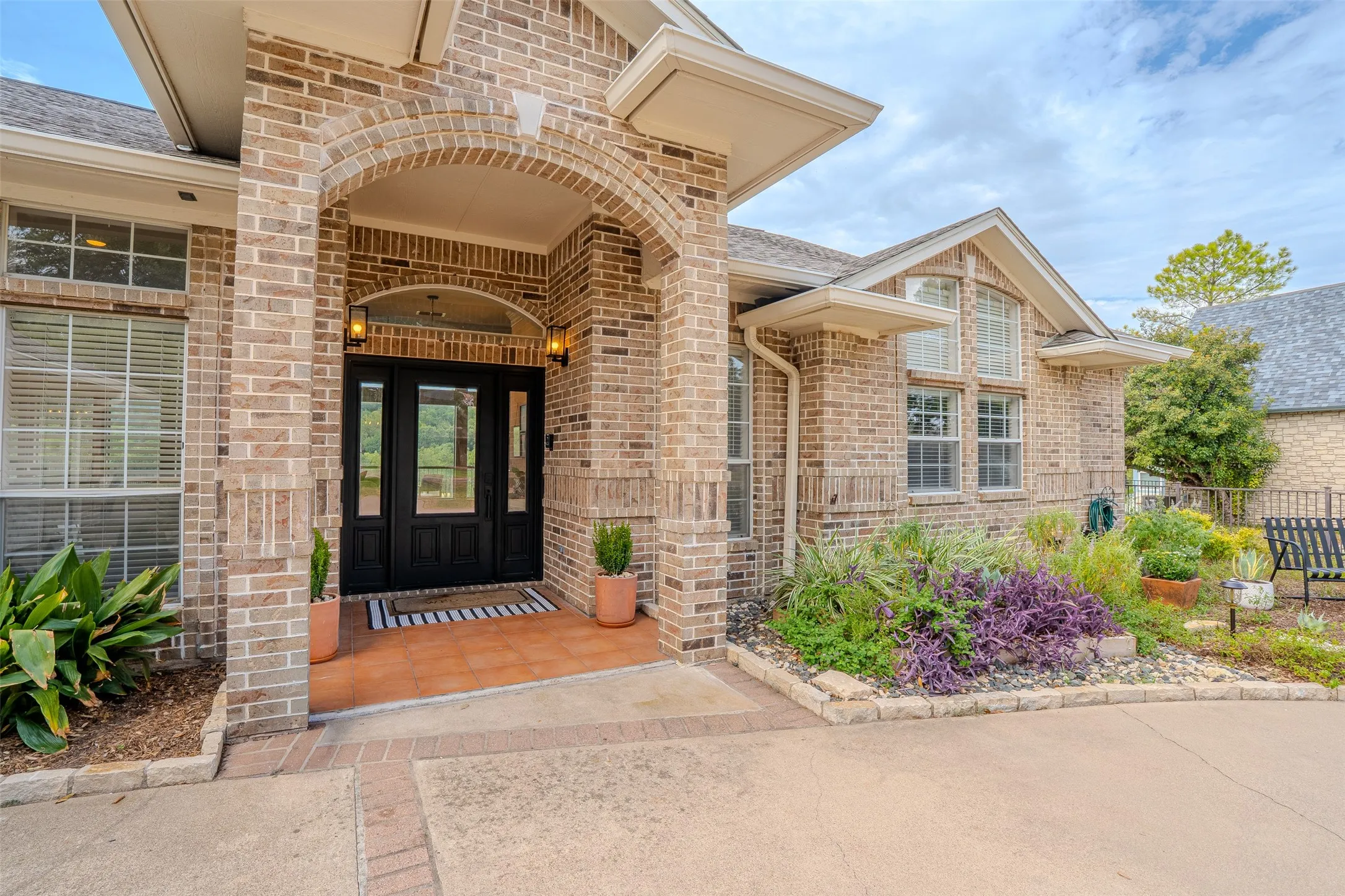 Brick facade residence featuring an arched portico entry with double doors, a tiled porch, and a landscaped garden bed