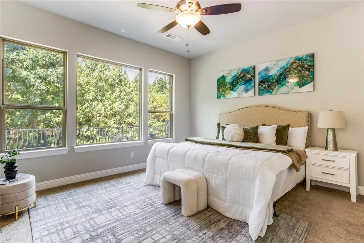 Spacious primary suite filled with natural light from three expansive windows overlooking lush greenery. Features neutral tones, plush carpeting, high ceilings, and a ceiling fan.