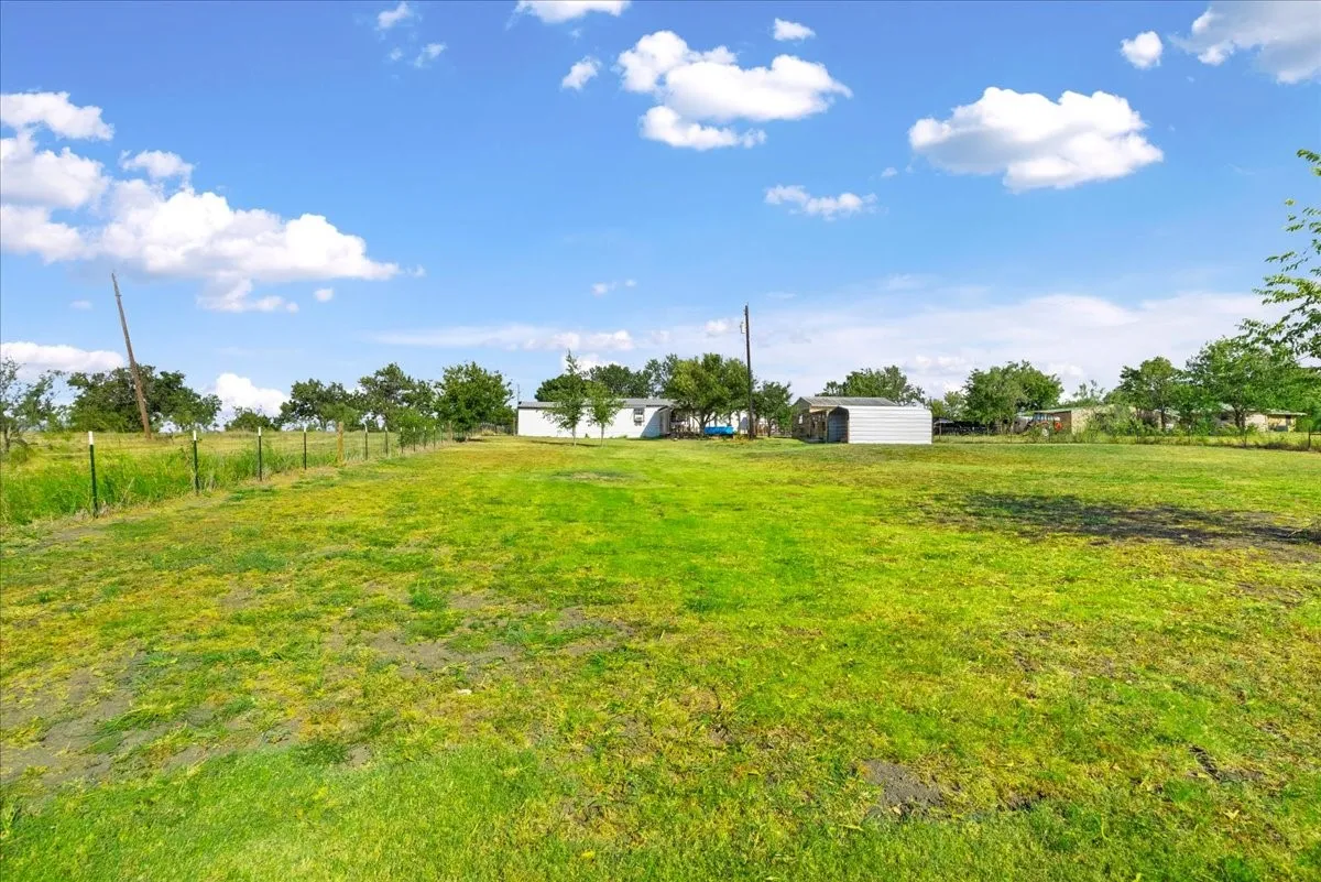 Expansive open field with a lush green lawn, partially fenced perimeter, and distant structures including a residence and an outbuilding