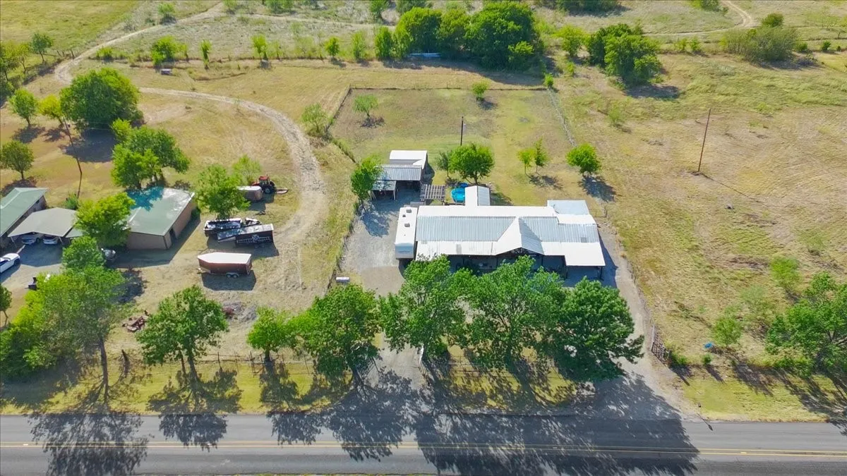 Aerial view of rural property featuring two modern structures with white metal roofing, expansive cleared grounds with mature tree coverage along the perimeter, established pasture areas, and direct road frontage