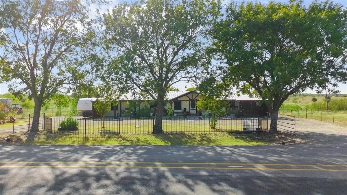Gated property entrance with a paved road frontage, mature trees, and a metal fence