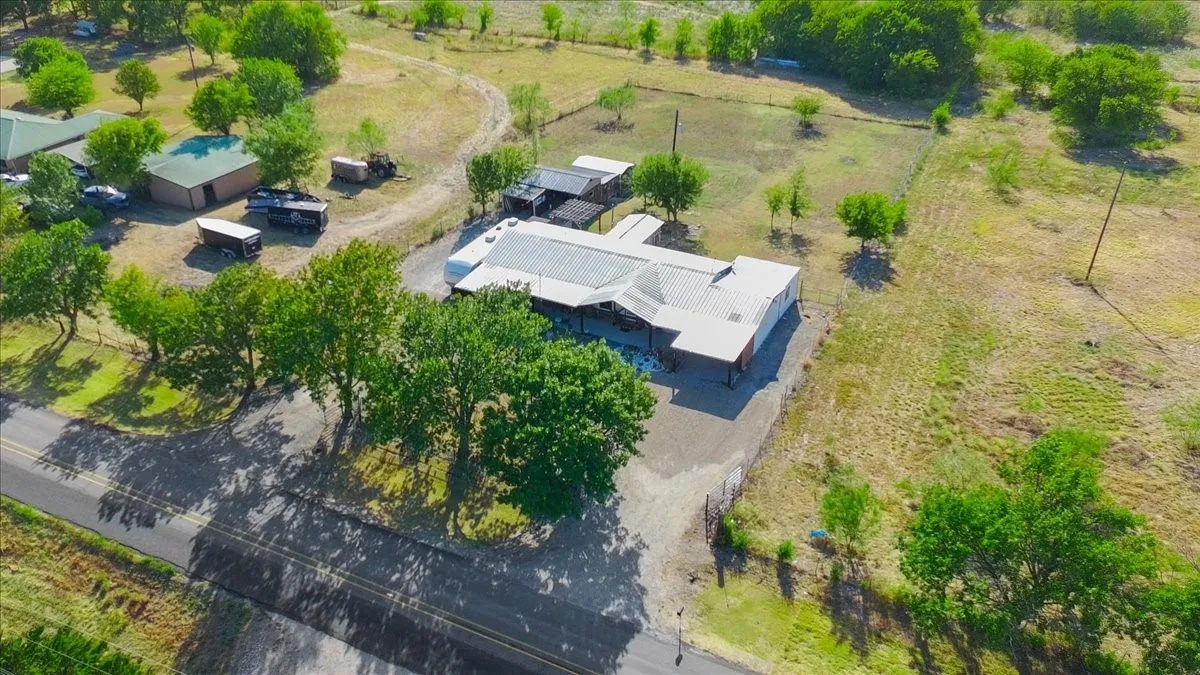 Main structure with metal roofing and covered outdoor areas, gravel driveway, surrounding green spaces, and mature trees