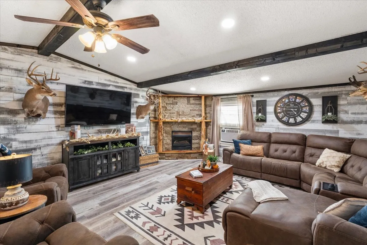 Expansive living area featuring a stone fireplace, wood-finish flooring, accent wall with horizontal paneling, decorative ceiling beams, and a ceiling fan with integrated lighting