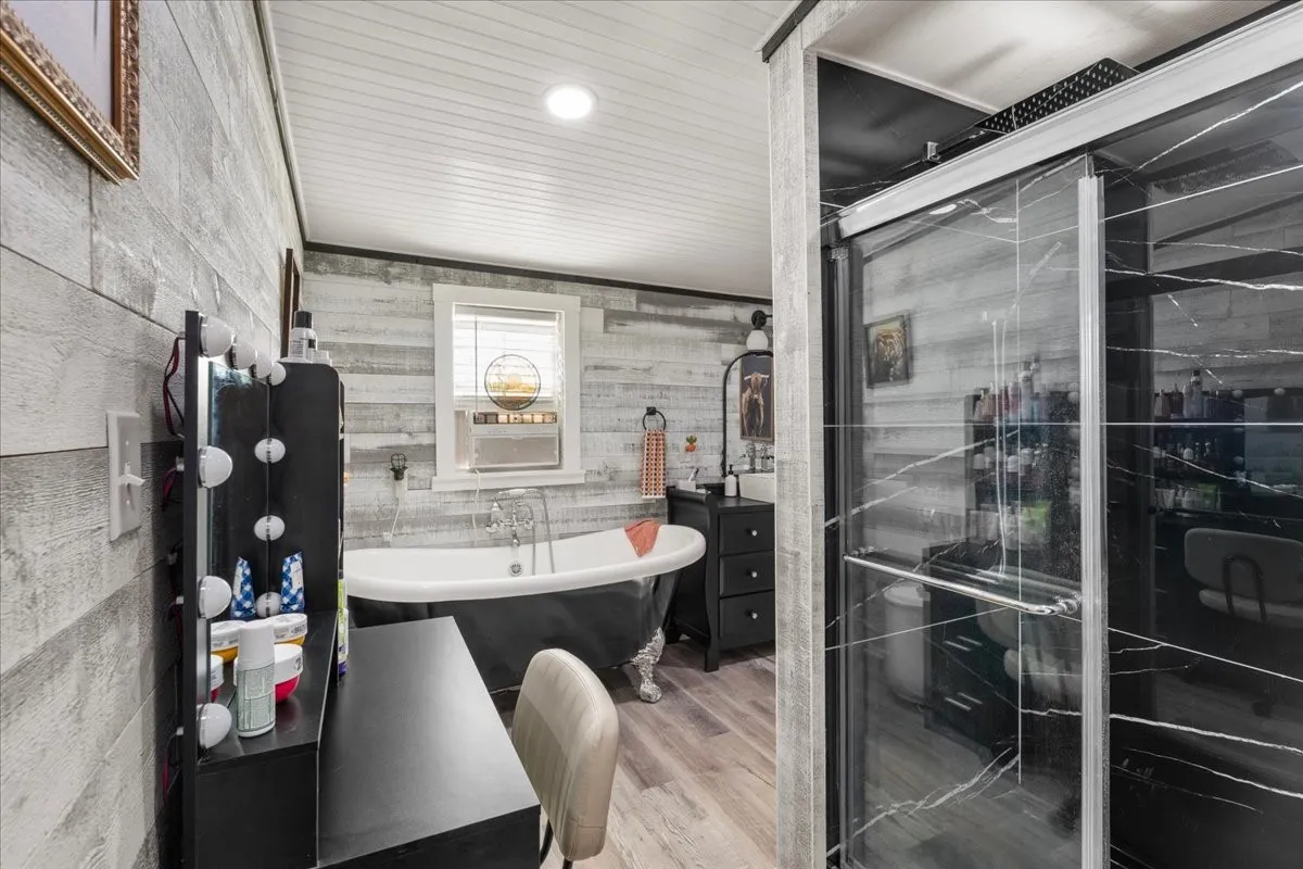 Freestanding clawfoot bathtub with a window above, glass-enclosed shower with dark stone-look tile, wood-finish flooring, and shiplap wall paneling