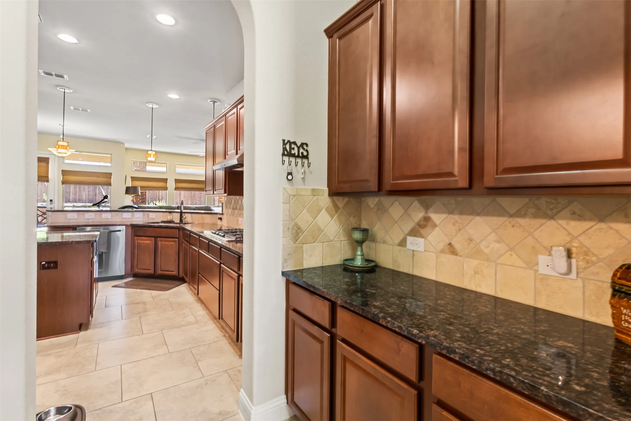 Arched entryway leading to a kitchen featuring wood-finish cabinetry, dark countertops, and a tiled backsplash