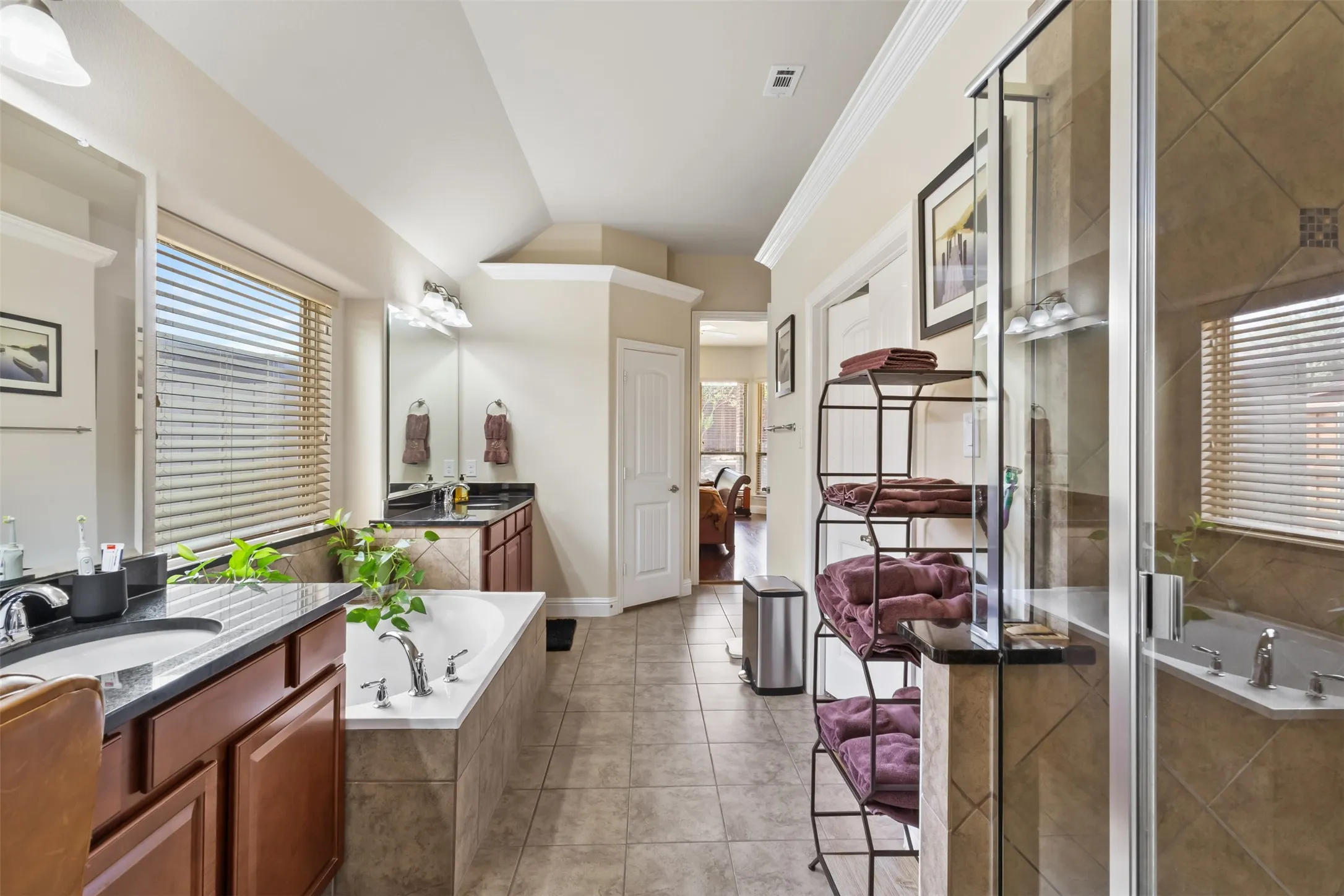 Dual vanities with dark stone countertops, built-in soaking tub, separate glass-enclosed shower, and tile flooring