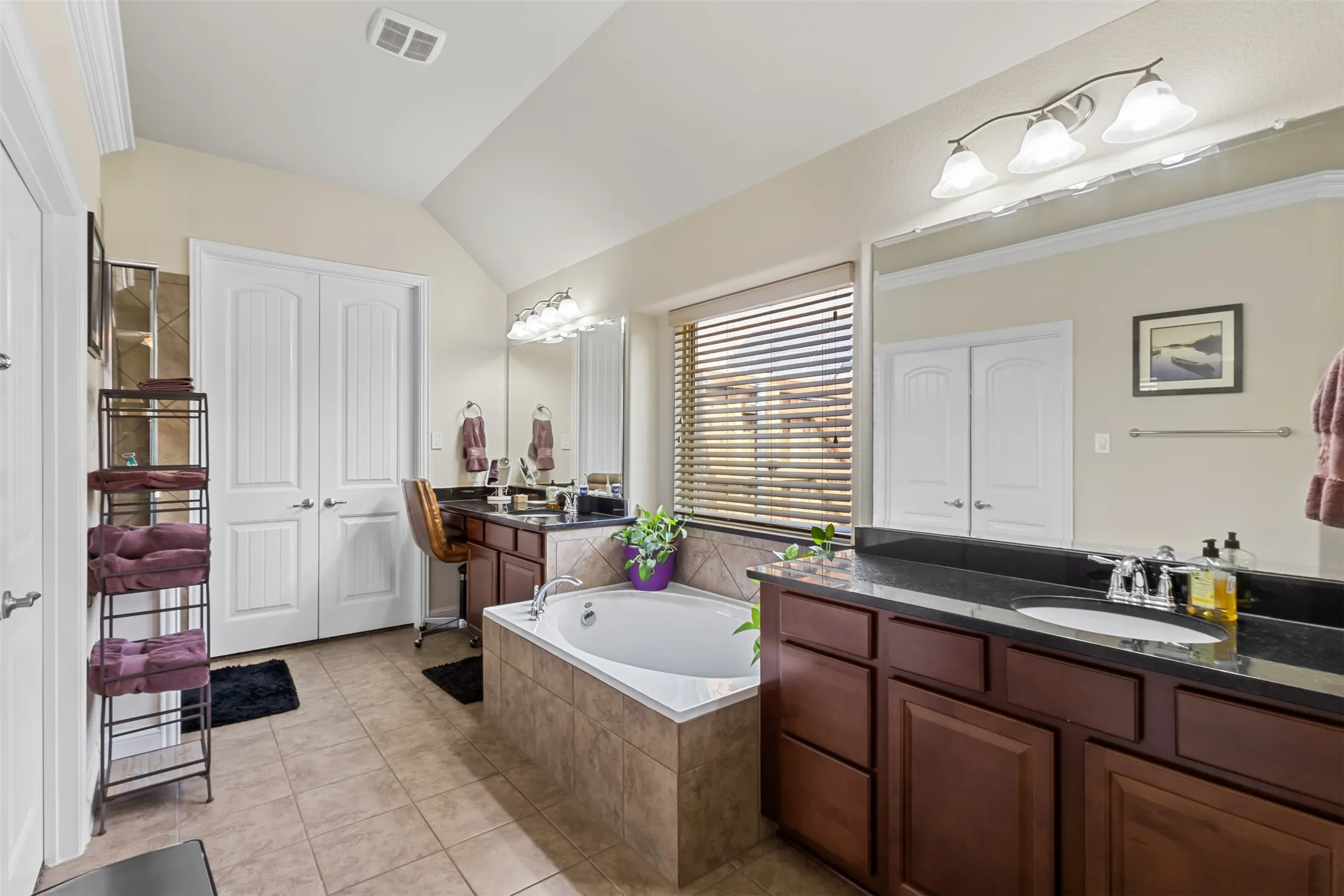 Vaulted ceiling, dual vanities with dark countertops, tiled flooring, built-in bathtub, and horizontal blinds