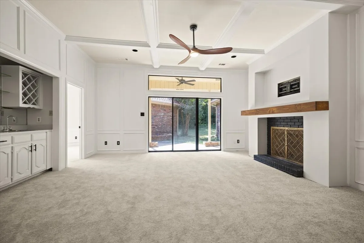 Coffered ceiling with a modern ceiling fan, built-in wet bar with white cabinetry, fireplace with a wood mantle, black brick hearth, and sliding glass doors