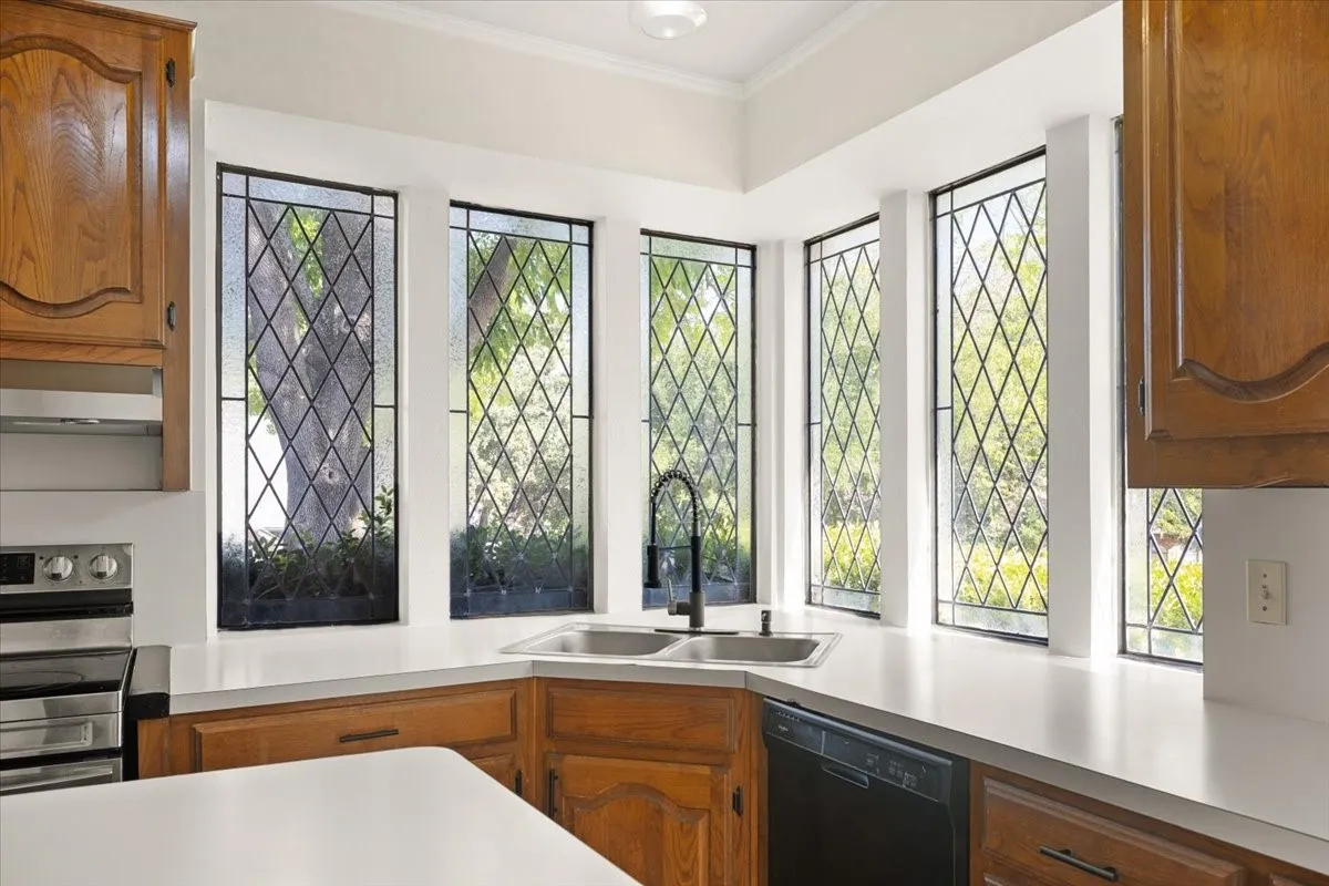 Bright corner kitchen featuring five leaded-glass casement windows with geometric lattice patterning, white quartz countertops, wood-finish cabinetry, and stainless steel built-in appliances including a wall-mounted oven unit