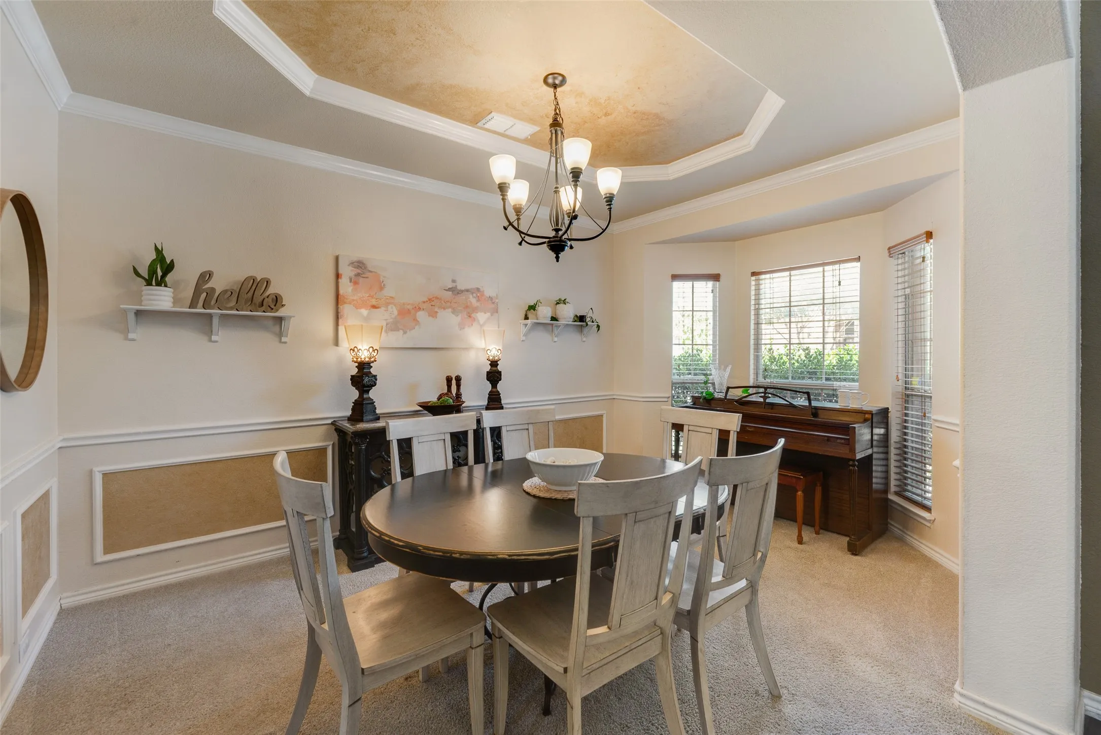 Tray ceiling with crown molding and central chandelier, bay window with multiple panes, wainscoting, and neutral-toned carpeting