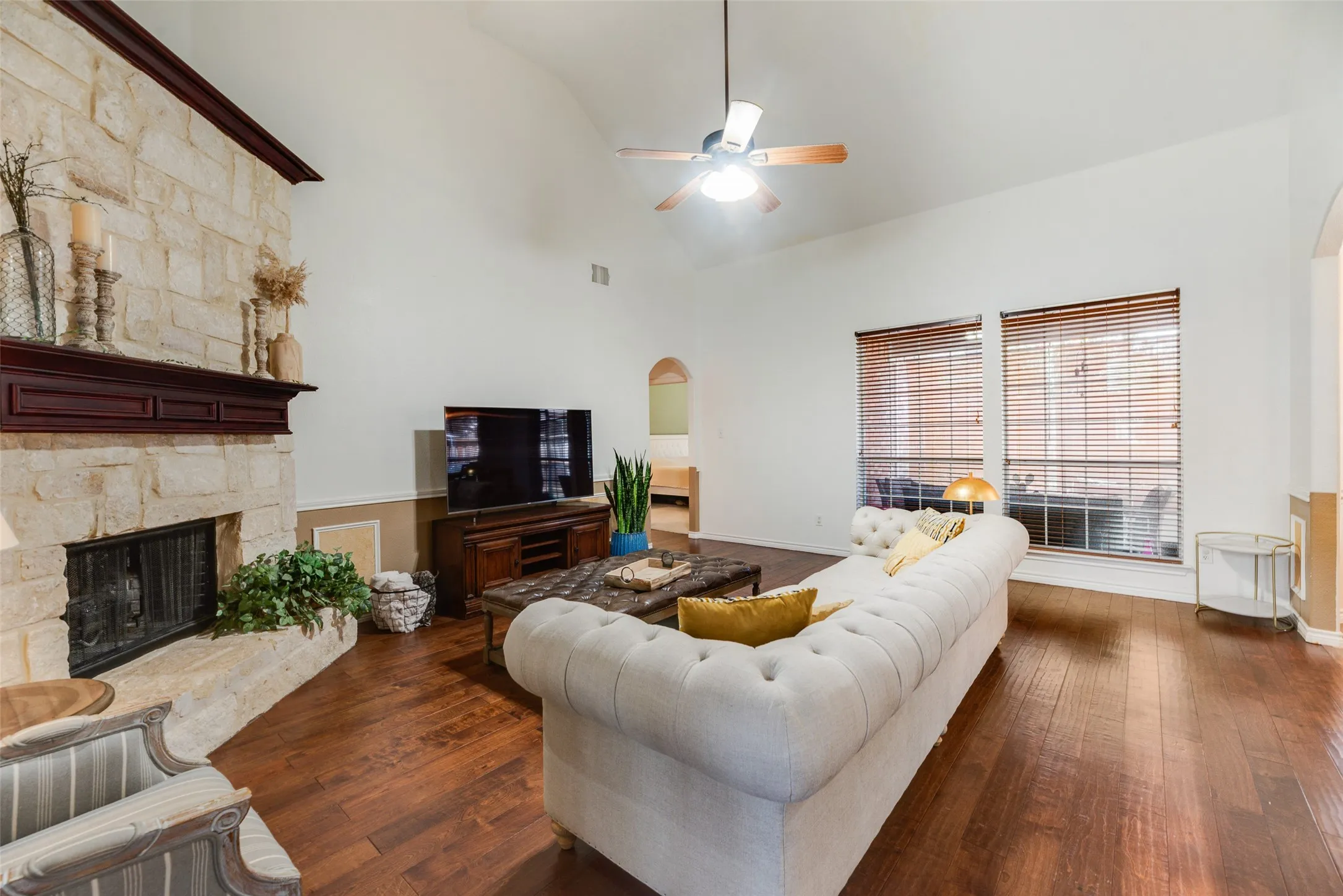 Stone-faced fireplace with a dark wood mantel, wood-finish flooring, vaulted ceiling, and dual windows with blinds