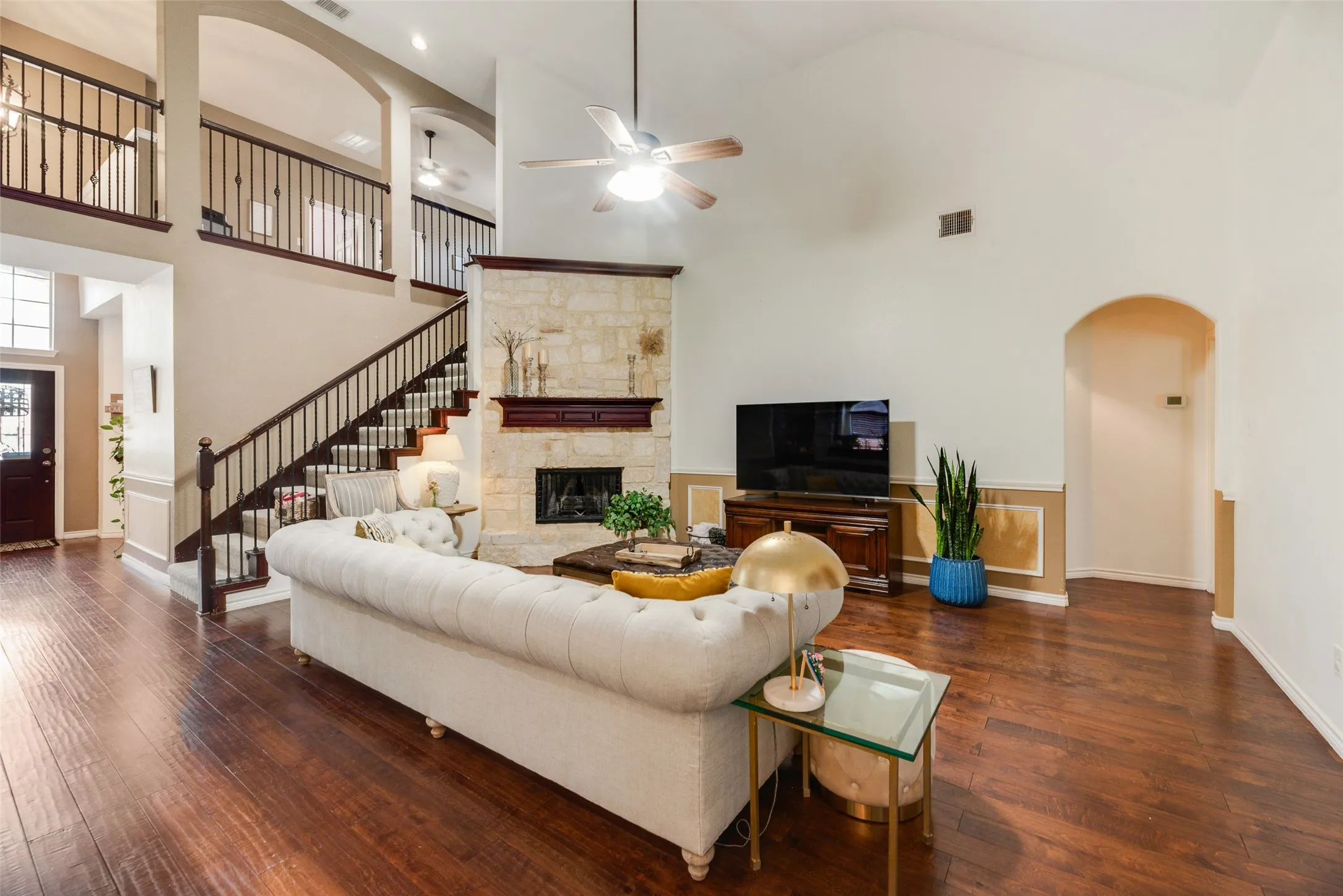 Hardwood flooring, vaulted ceilings, stone fireplace, open staircase with wrought iron balusters, and a ceiling fan