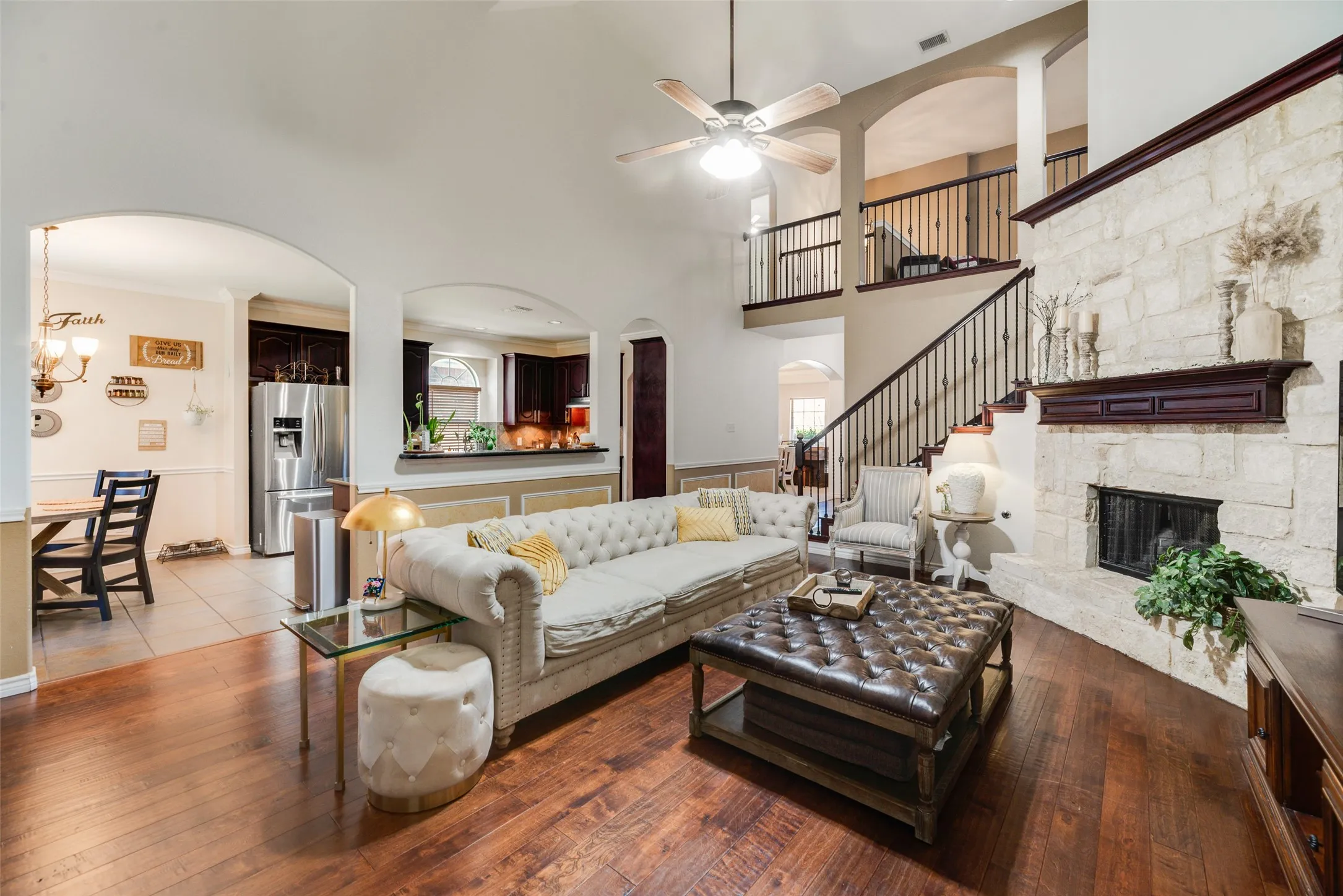 Hardwood-finish flooring, stone-clad fireplace with dark wood mantel, high vaulted ceiling with fan, open-concept design with arched doorways, and a dark wood and wrought iron staircase