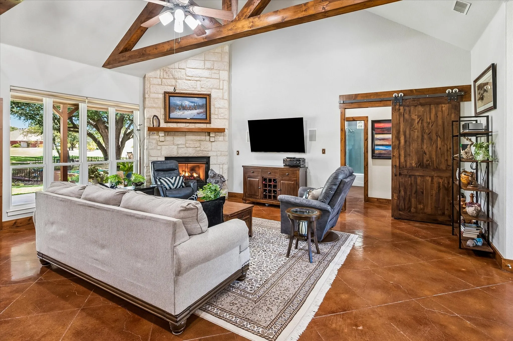 Vaulted ceiling with exposed wood beams, stone fireplace, large windows, stained concrete flooring, and a sliding barn door