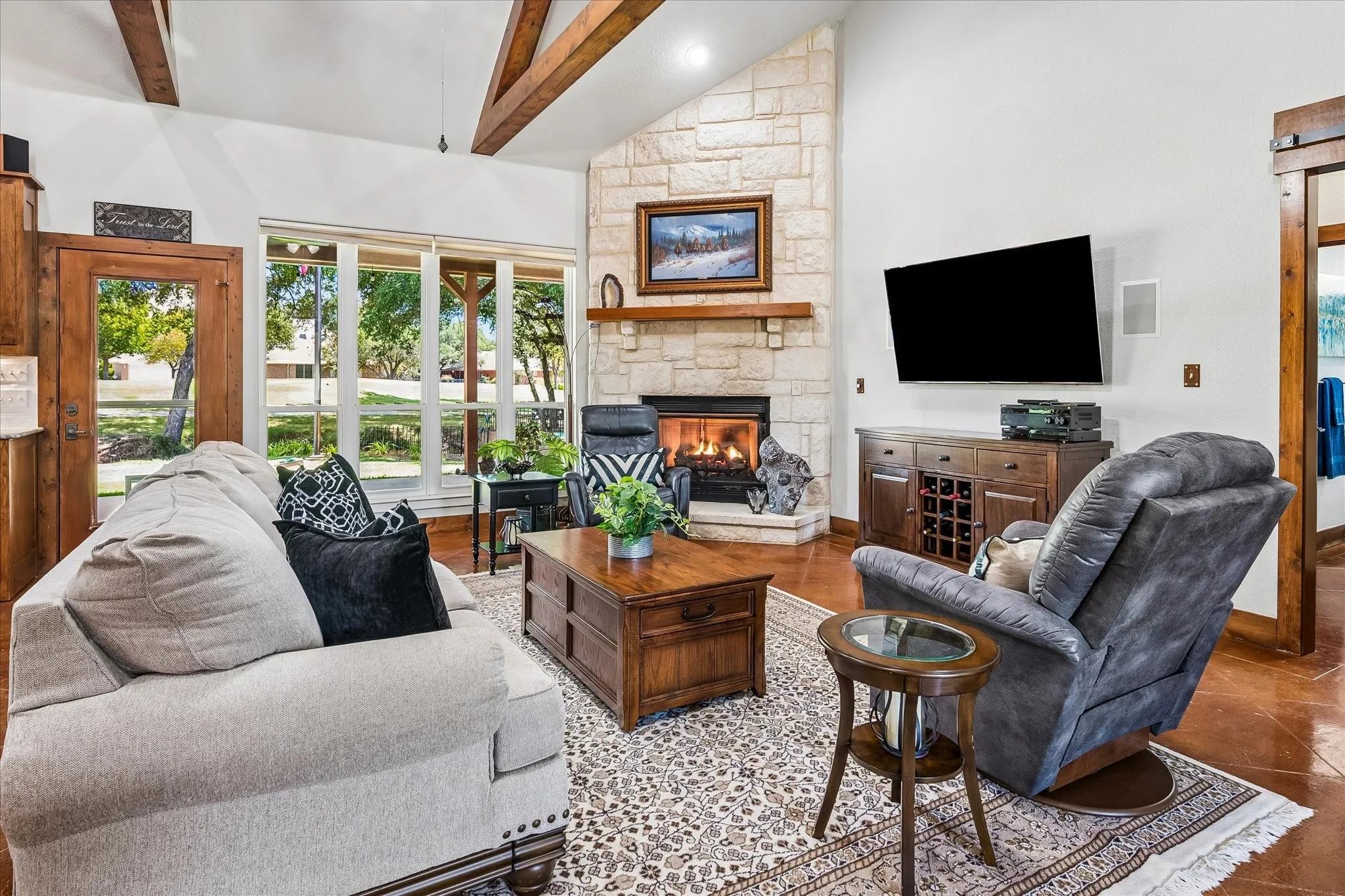Vaulted ceiling with exposed wood beams, stone-faced fireplace with wood mantle, large picture windows, wood-finish flooring, and a barn door entry
