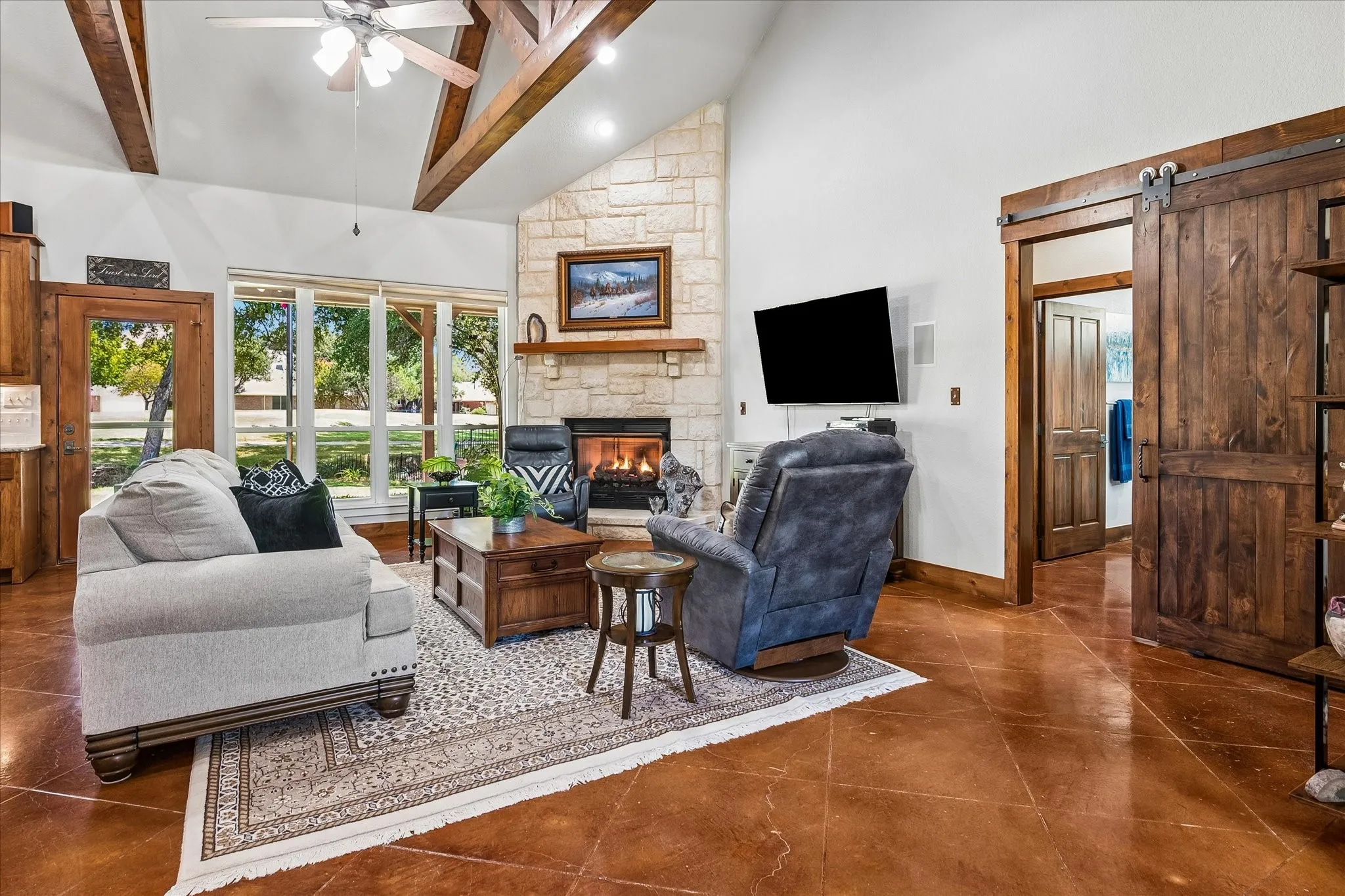 Vaulted ceiling with exposed wood beams, stone fireplace, large windows, stained concrete flooring, and a wood barn door