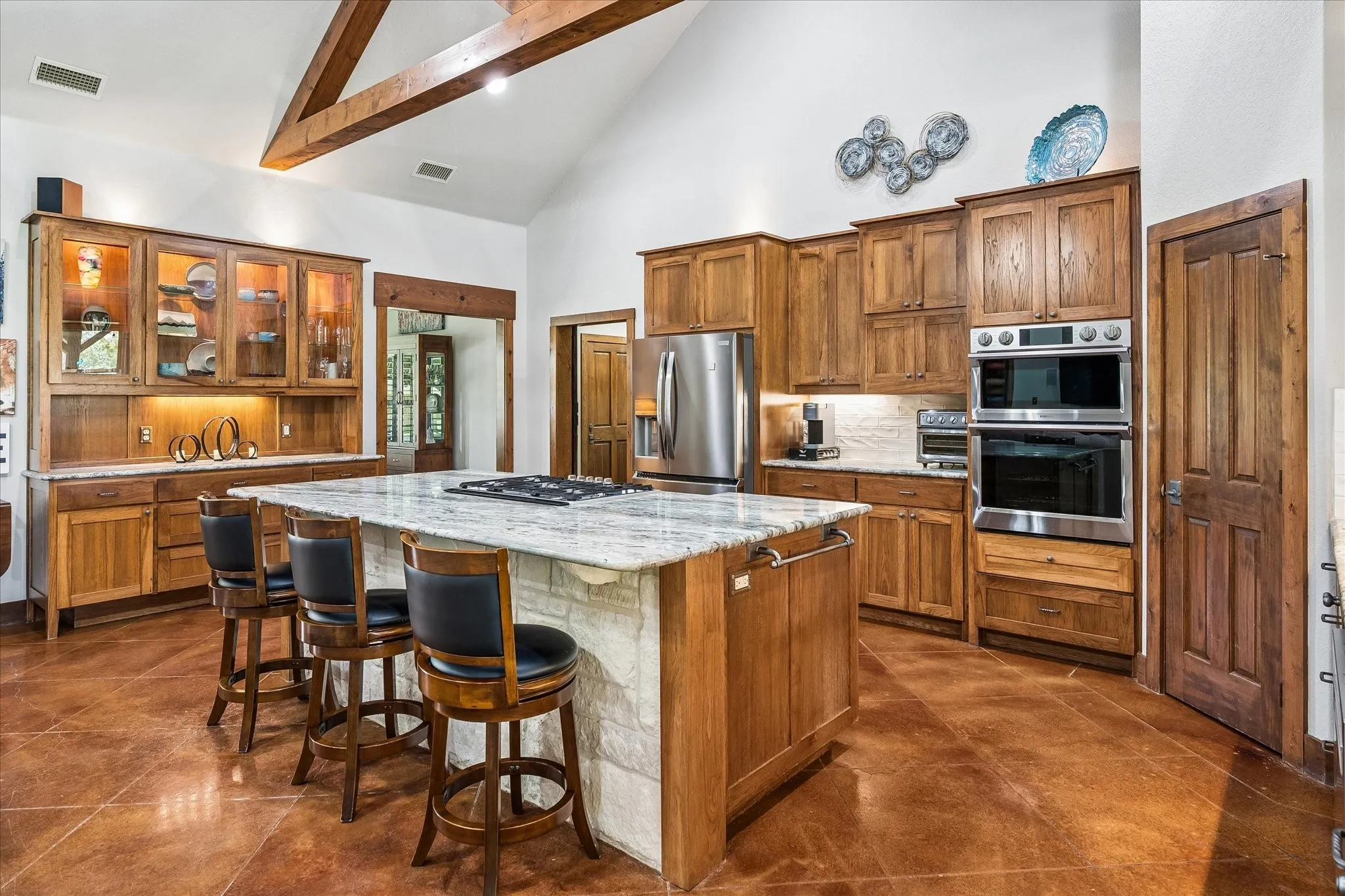 Vaulted ceiling with exposed wood beams, wood-finish flooring, custom wood cabinetry, stainless steel appliances, and a central island with a cooktop
