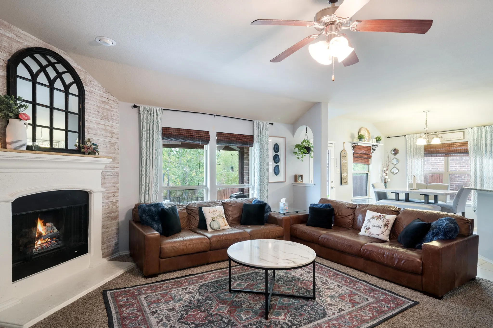 Living area featuring a fireplace with a mantel, a ceiling fan, and large windows with blinds and drapes