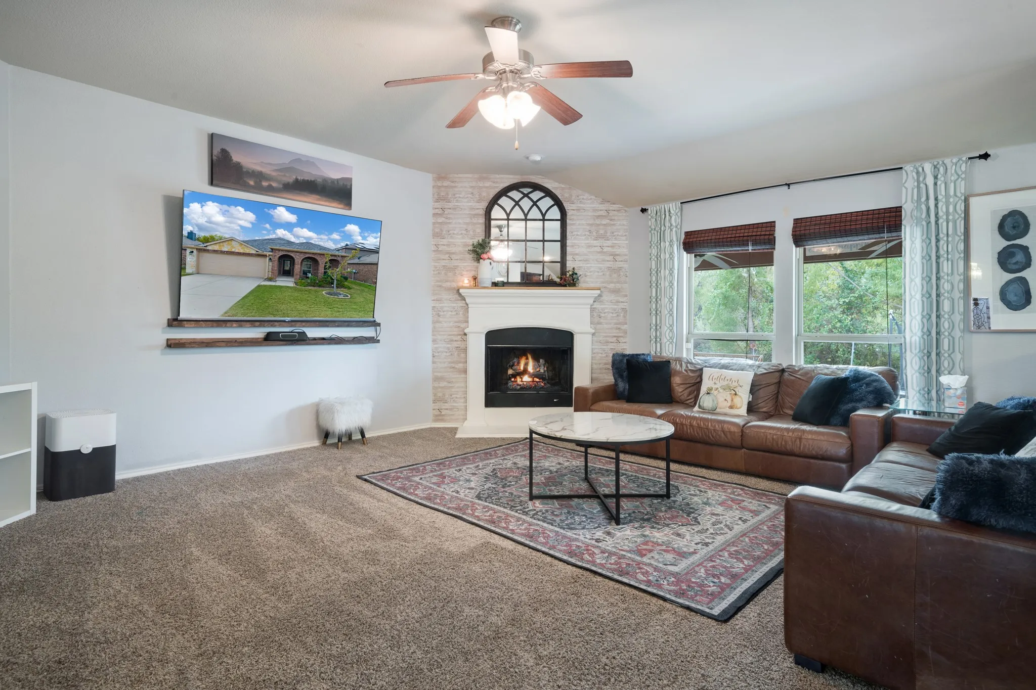 Carpeted living area featuring a central ceiling fan, a fireplace with a white mantel and distressed brick surround, and multiple windows