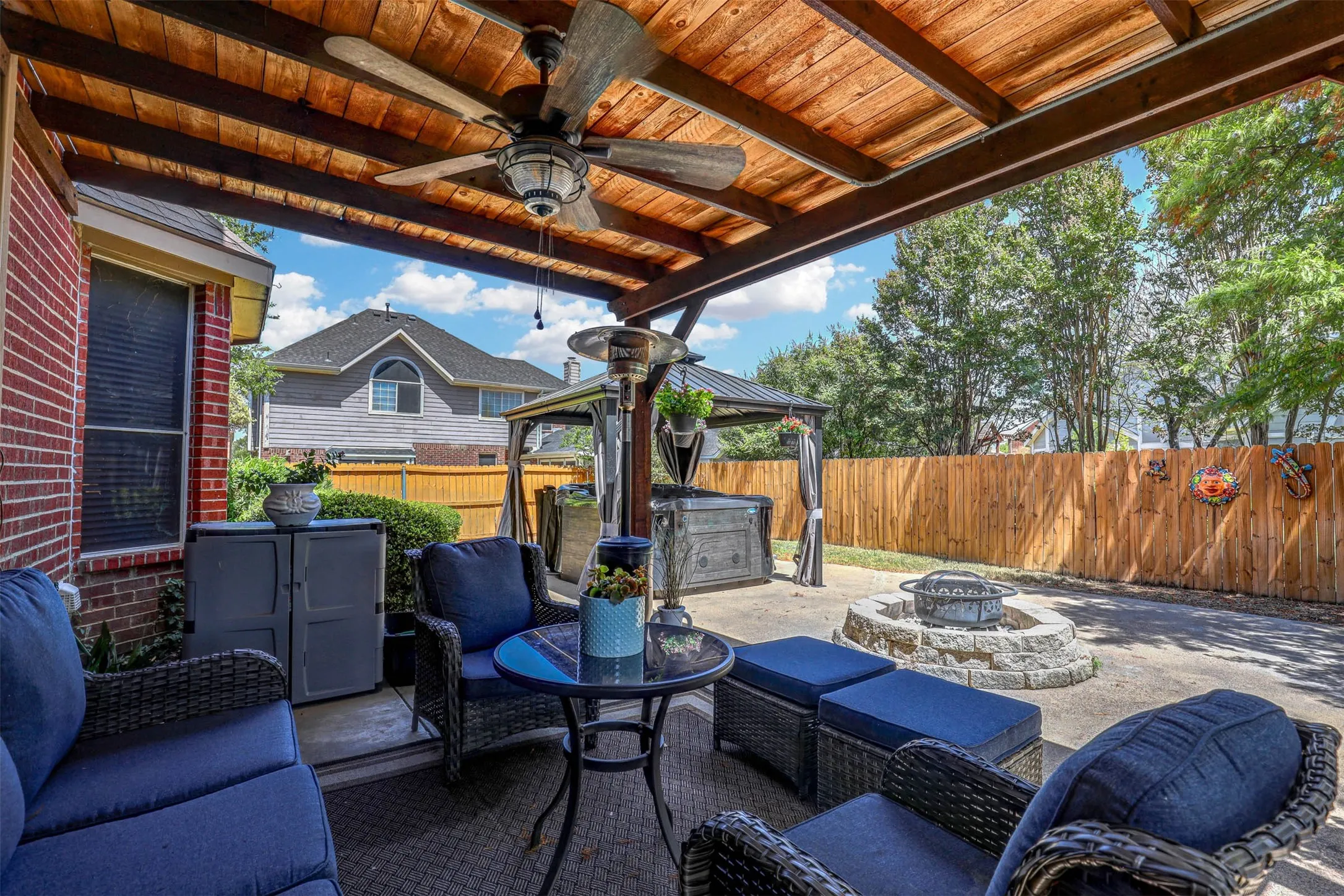 The covered patio offers a serene vantage point over the tree-lined backyard with a clear view of the fire pit, hot tub, and spacious open lawn.