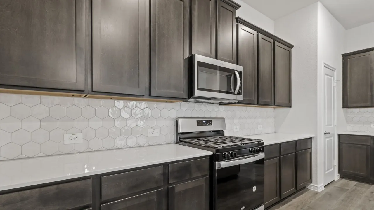 Dark wood-finish cabinetry, stainless steel microwave, gas range, white hexagon tile backsplash, and light-toned countertops