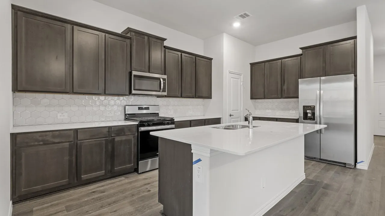 Spacious kitchen featuring dark wood-finish cabinetry, stainless steel appliances, hexagon tile backsplash, light-colored countertops, and a kitchen island with an integrated sink