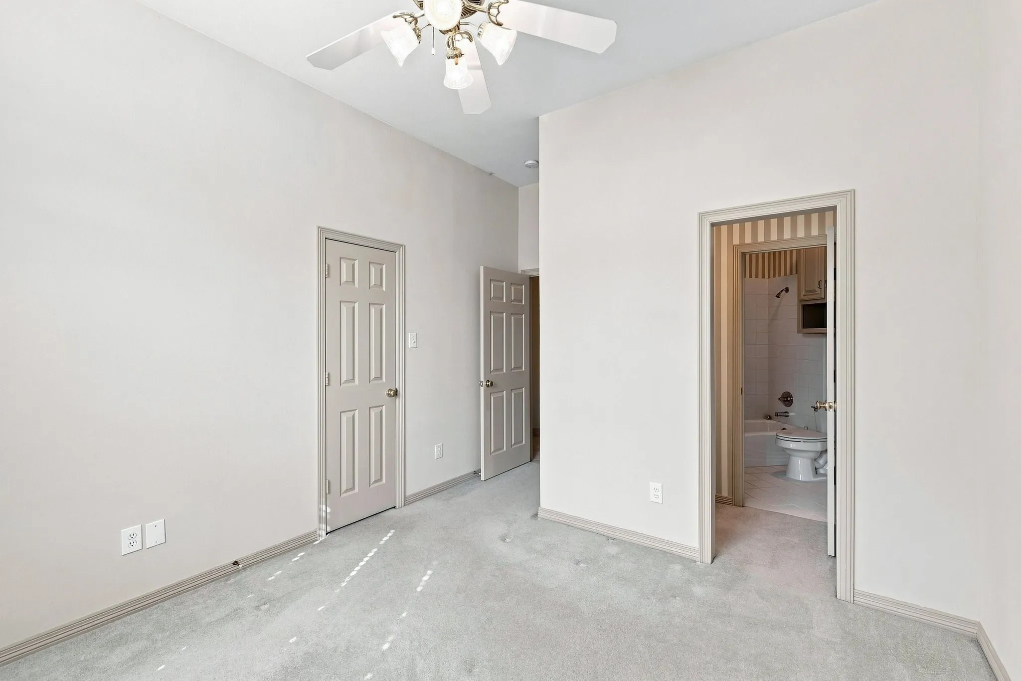 Carpeted room with a ceiling fan, multiple paneled doors, and access to a bathroom featuring a tub and built-in cabinetry