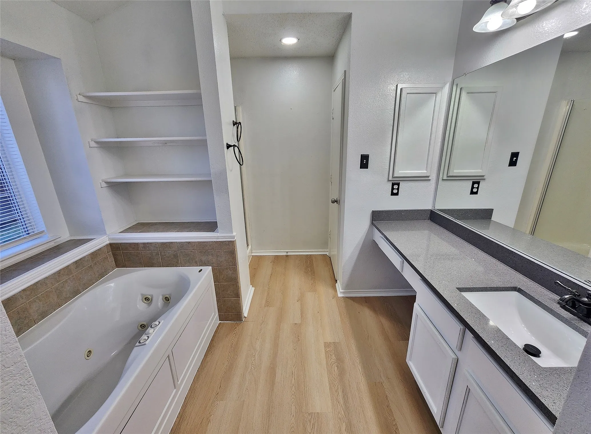 Jetted soaking tub, built-in shelving, wood-finish flooring, dual vanity with undermount sink, and stone-finish countertop
