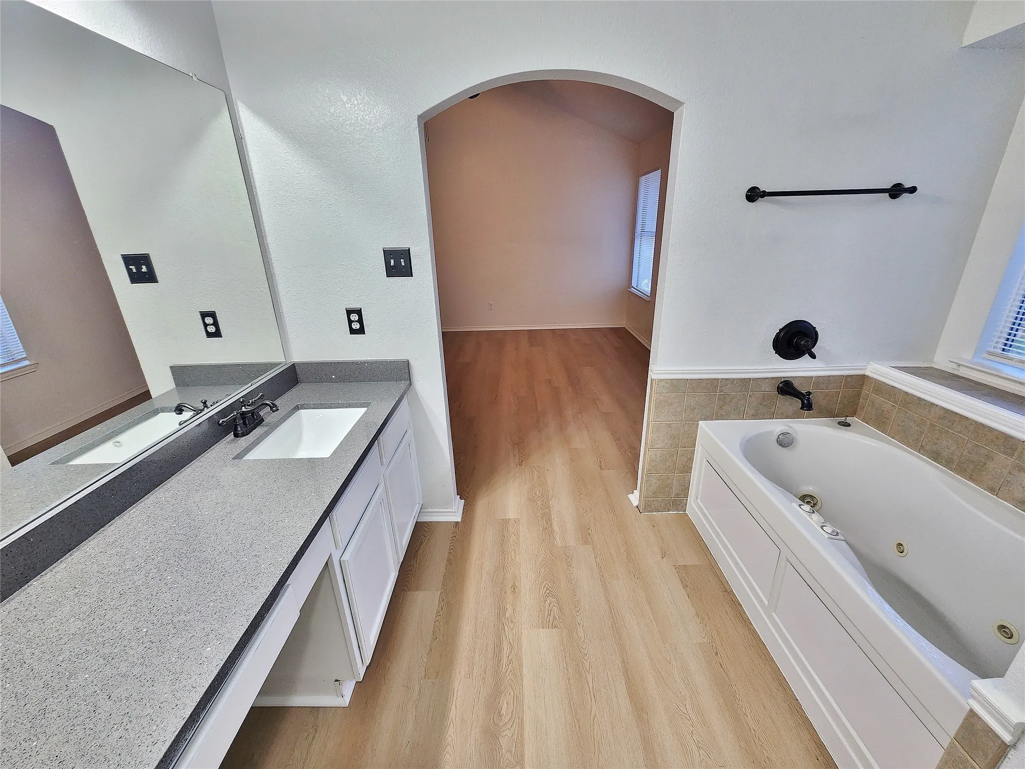 Dual vanity with integrated sinks, spacious jetted tub, wood-finish flooring, and an arched entry
