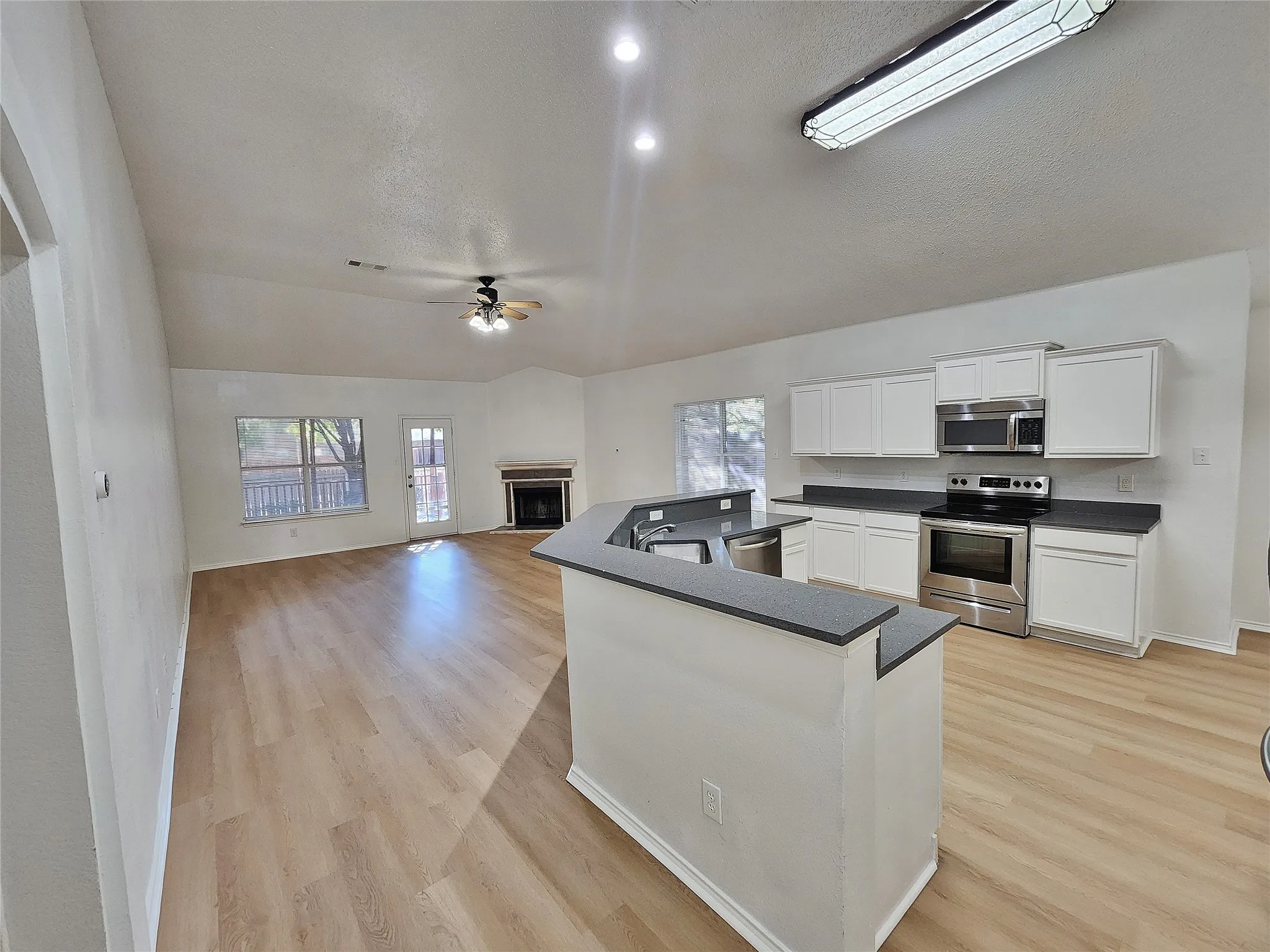Open-concept living space featuring wood-finish flooring, a fireplace, white kitchen cabinetry, stainless steel appliances, and dark countertops